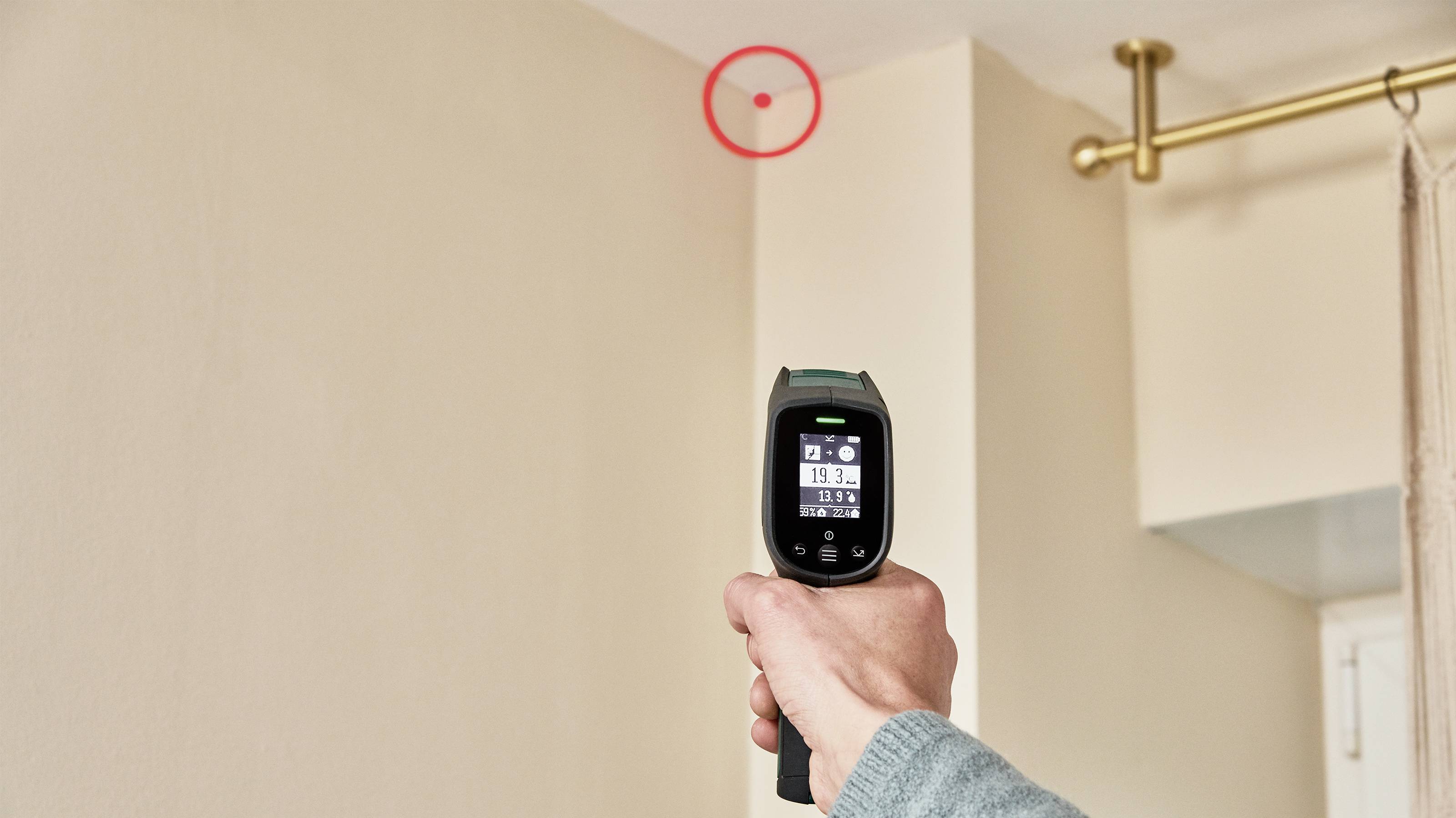Une personne pointe un thermomètre infrarouge vers un marquage rouge sur le mur pour mesurer la température.