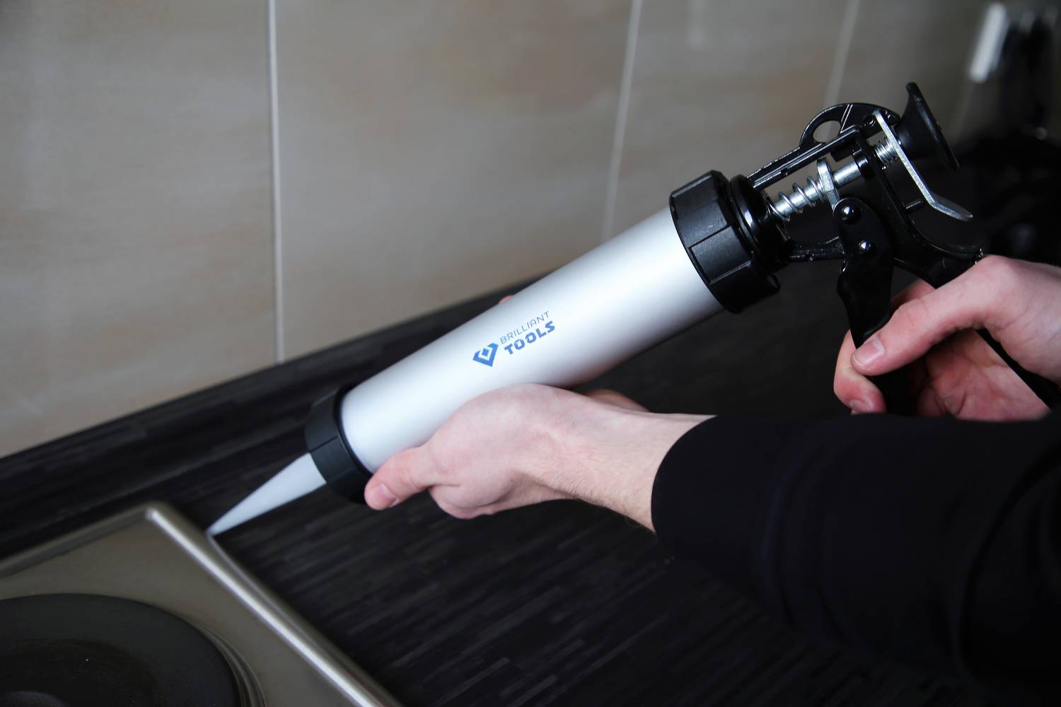 Une personne tient un pistolet à cartouche et applique un mastic d'étanchéité sur le joint entre la plaque de cuisson et le plan de travail.