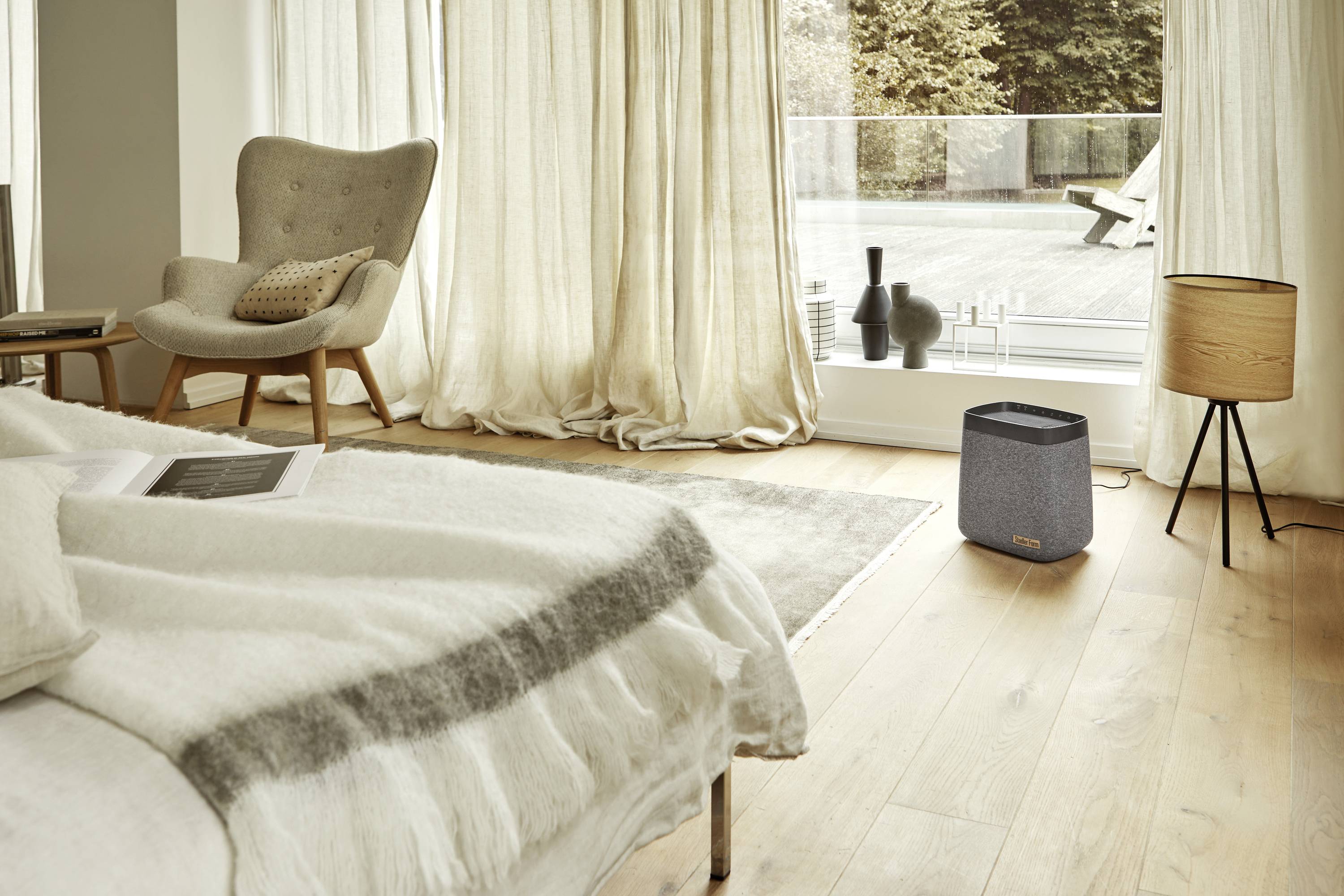 Une chambre lumineuse avec un sol en bois, un fauteuil gris et un haut-parleur posé sur le sol. De grandes fenêtres donnent sur une terrasse avec un transat.