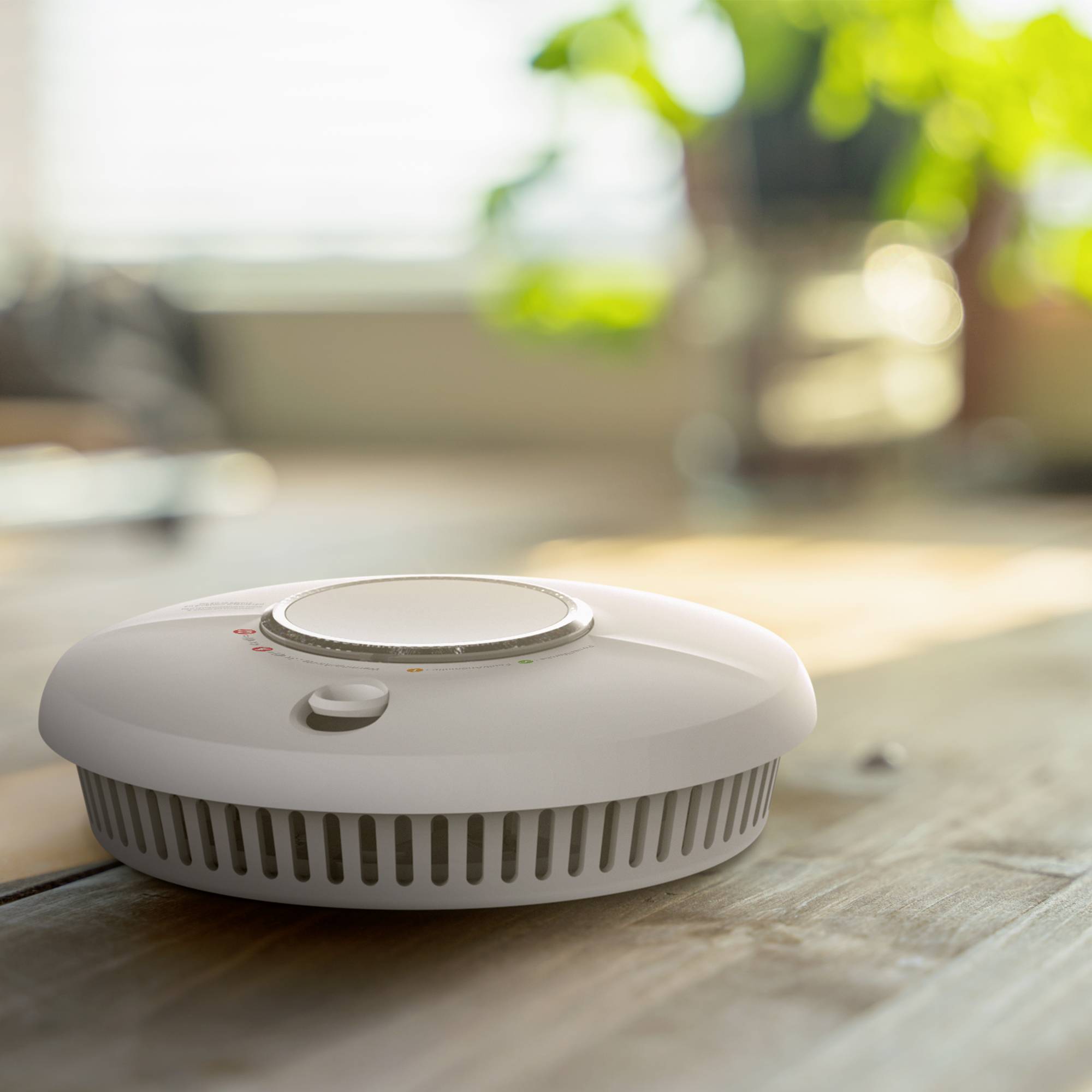 Un détecteur de fumée avec bouton de test et voyants LED indiquant l'état de fonctionnement, une plante en arrière-plan.