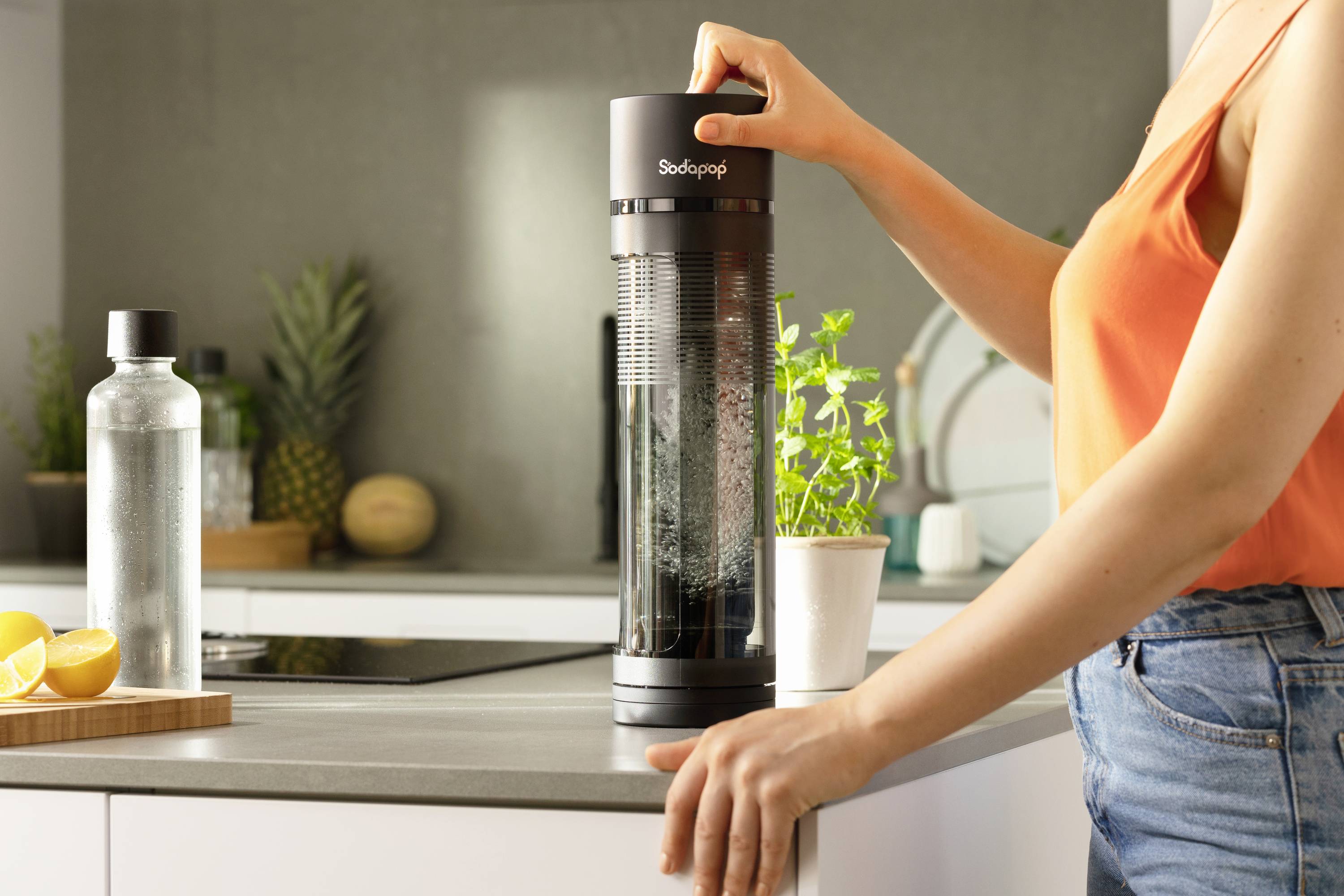 Une personne utilise une machine à gazéifier l'eau sur un plan de travail de cuisine. Des ustensiles de cuisine et une plante sont visibles en arrière-plan.