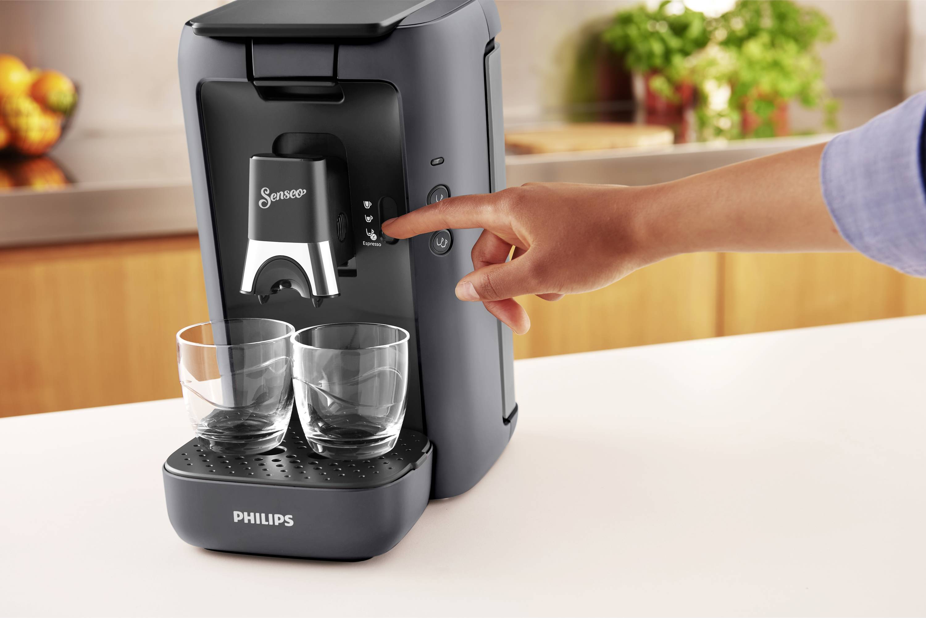 Une personne actionne une machine à café avec deux verres vides placés en dessous. En arrière-plan, on peut voir un plan de travail de cuisine avec des fruits.