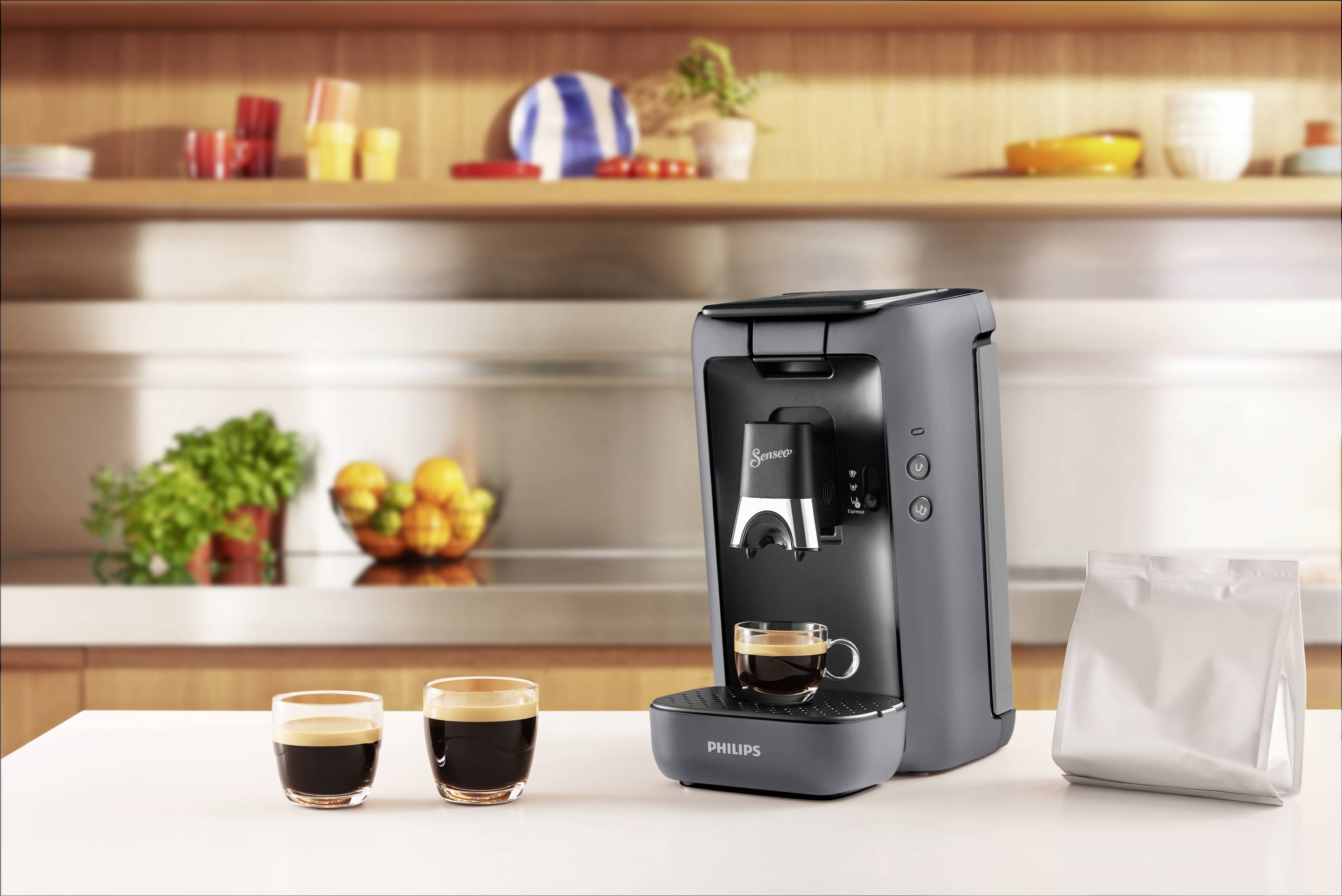 Une machine à café grise est posée sur un plan de travail de cuisine avec deux tasses de café remplies et un sachet à proximité. En arrière-plan, on aperçoit des étagères avec de la vaisselle et des fruits.