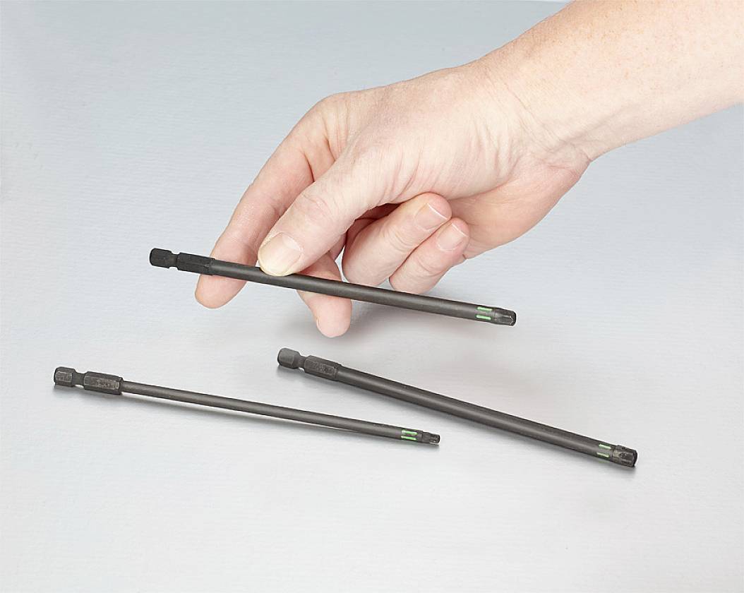 Une main tient une fine tige noire, deux autres tiges sont posées sur une surface. Les tiges ressemblent à des outils ou des pièces accessoires.