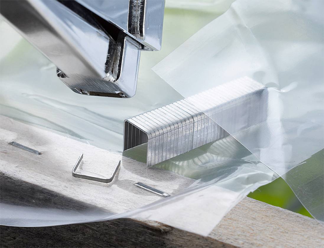 Une photo en gros plan montre un agrafeur fixant un agrafe métallique à travers un film plastique transparent sur du bois.