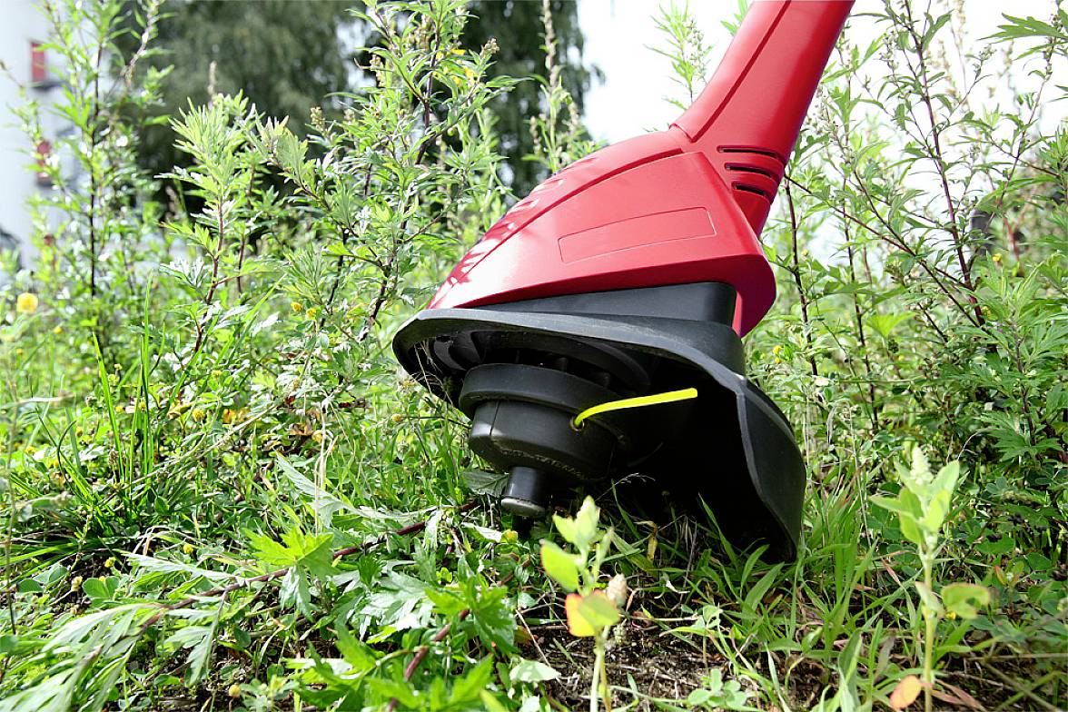 Un coupe-bordure rouge coupe l'herbe haute et les mauvaises herbes dans un jardin. Des buissons et des arbres sont visibles en arrière-plan.