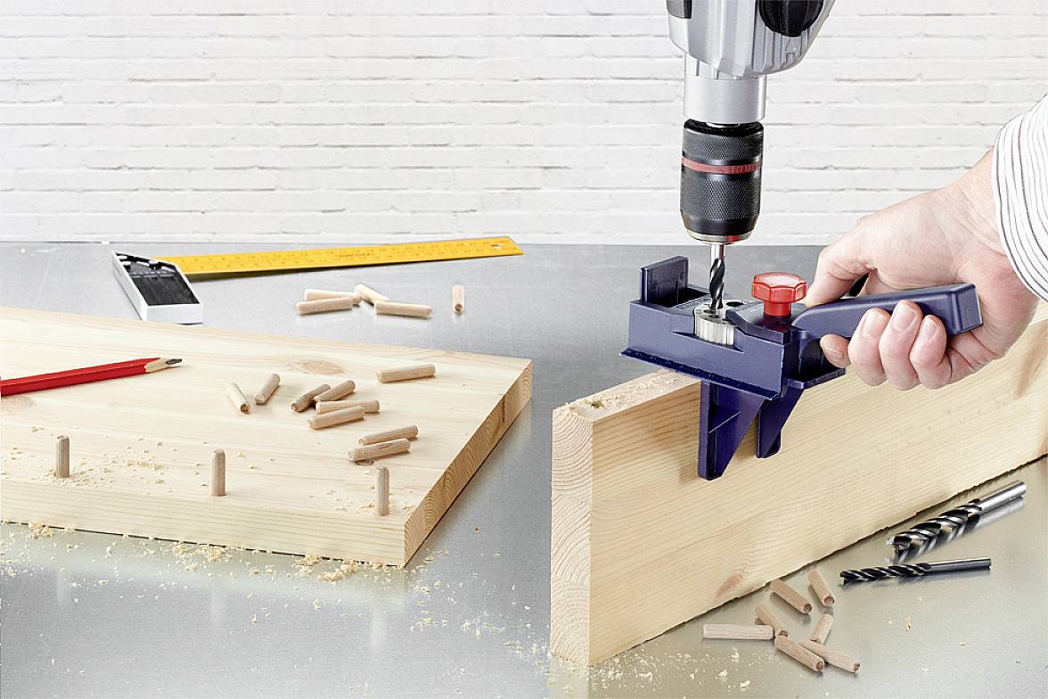 Un artisan fore des trous précis dans une planche de bois à l'aide d'un support de perceuse. Sur la table sont disposés des chevilles et des forets comme matériaux de travail.