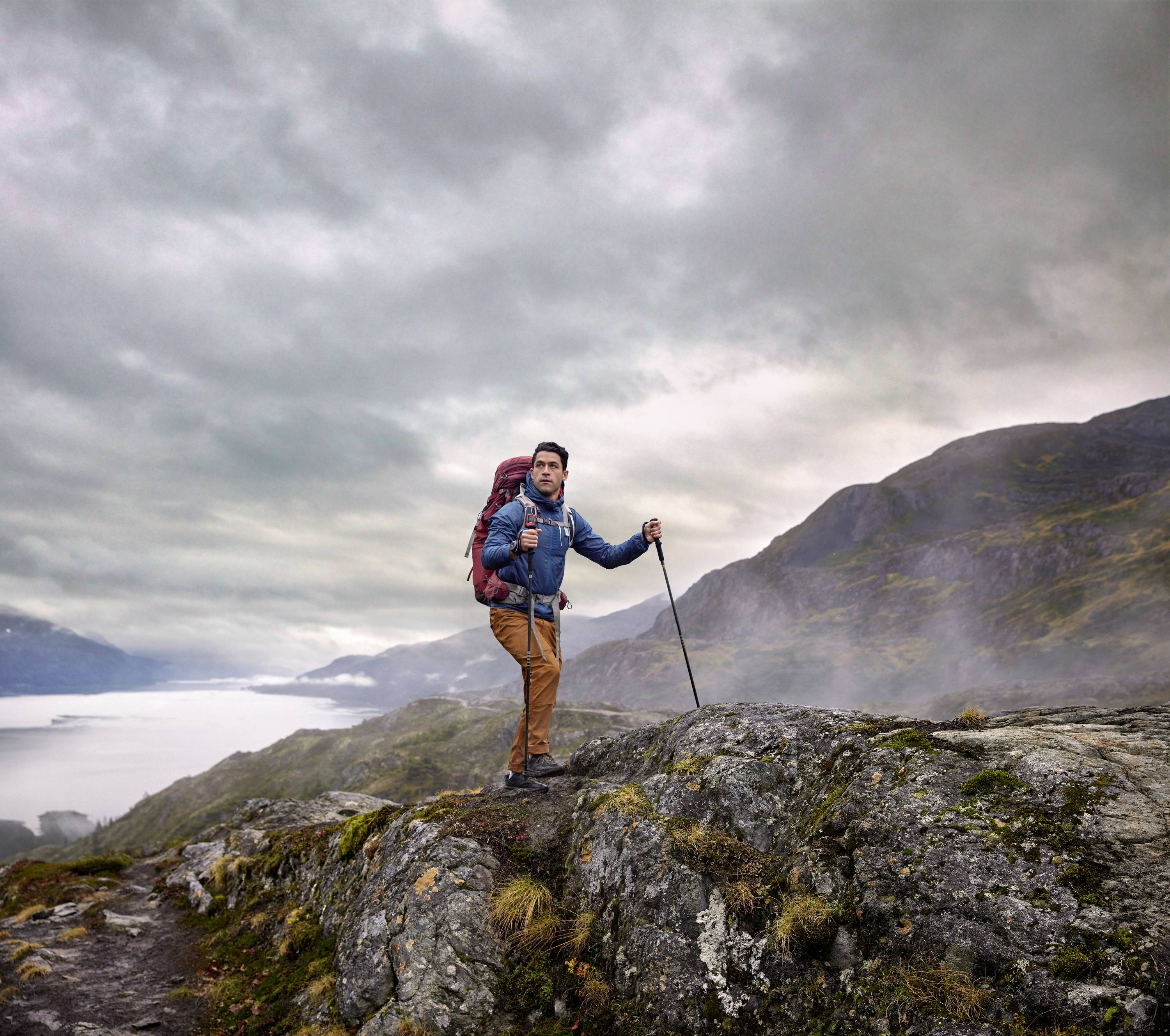 Une personne marche avec un sac à dos et une canne de randonnée sur un sentier rocailleux dans un paysage montagneux sous un ciel nuageux.