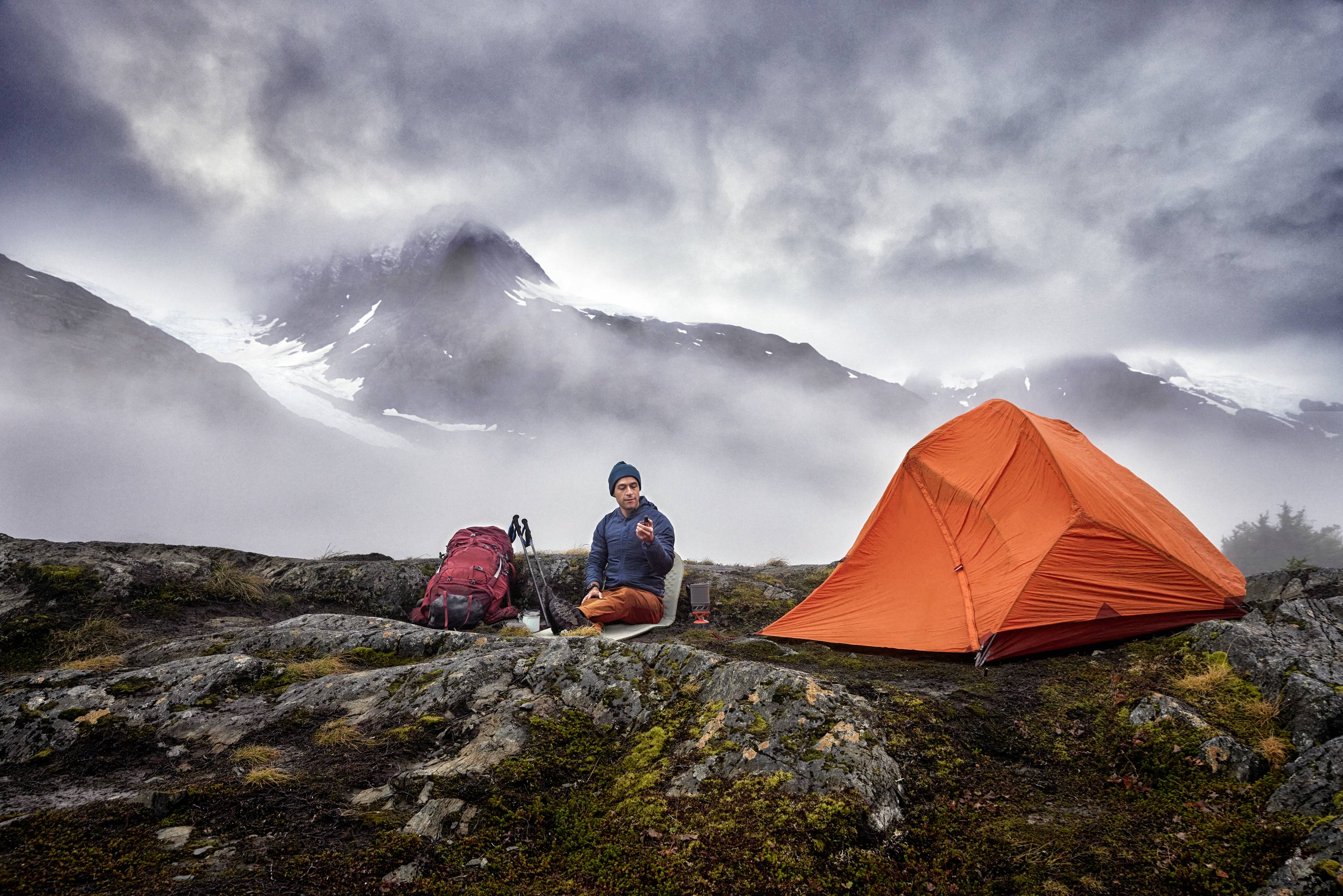 Une personne est assise à côté d'une tente orange dans un paysage montagneux et brumeux. Un sac à dos est posé à proximité.