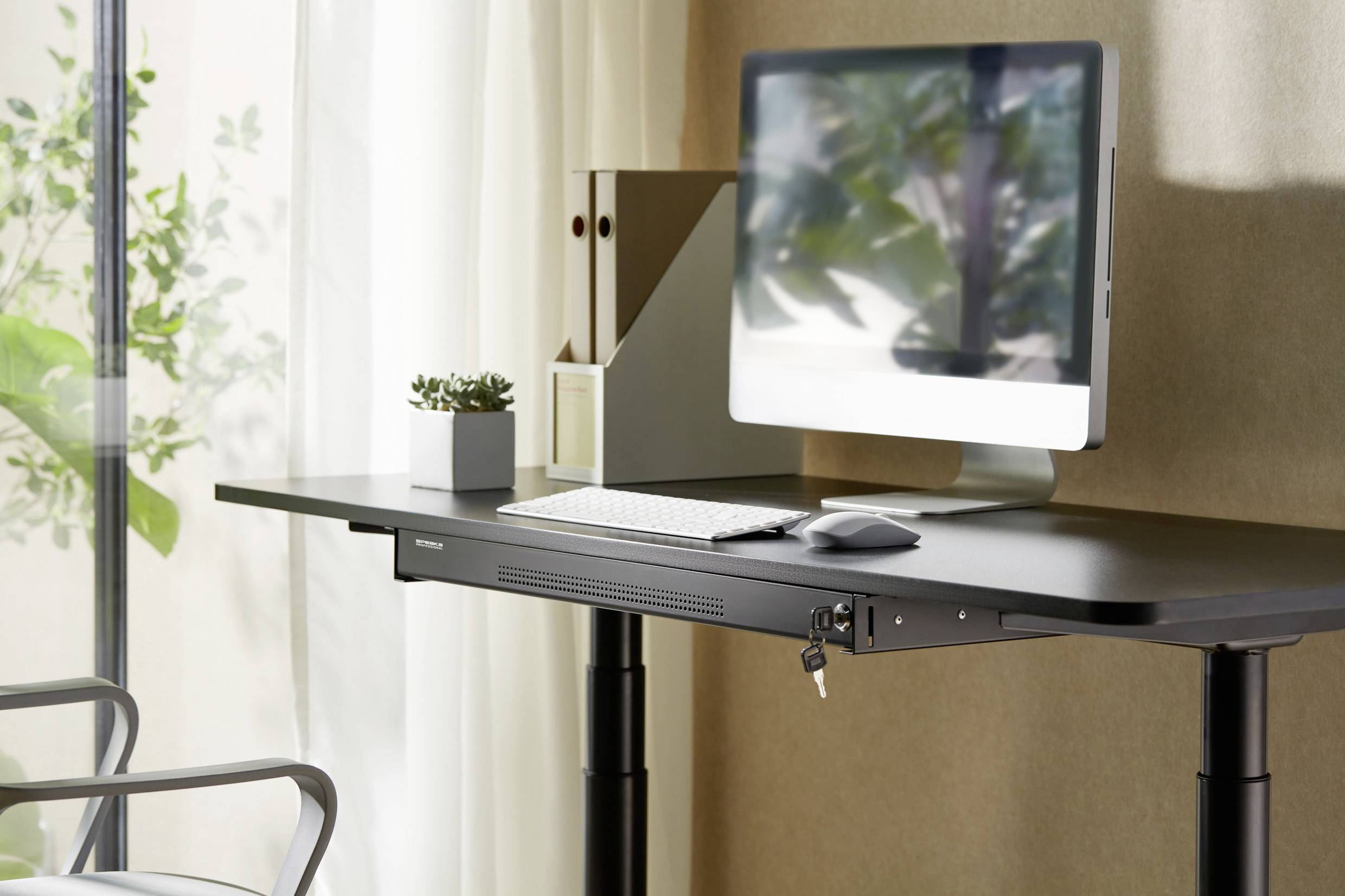 Un bureau à hauteur réglable avec un moniteur, un clavier, une souris et des plantes. À l'arrière-plan, des fenêtres avec des rideaux.
