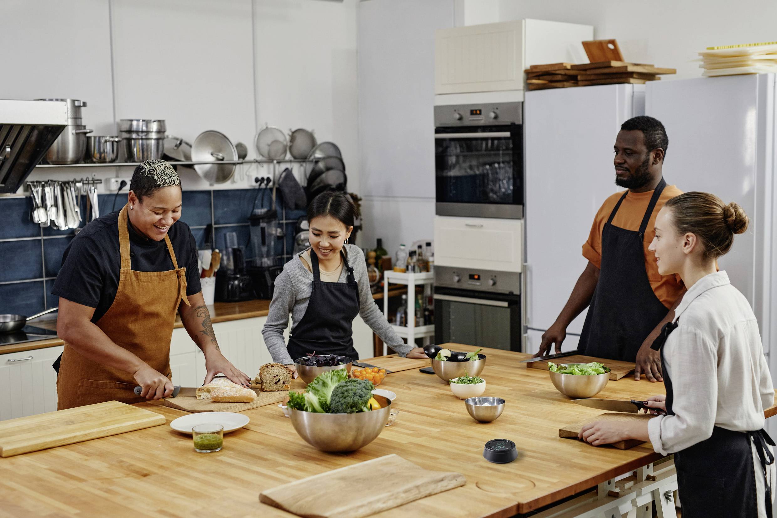 Quatre personnes portant des tabliers sont rassemblées autour d'une table de cuisine et préparent ensemble des légumes. Elles rient et se regardent.