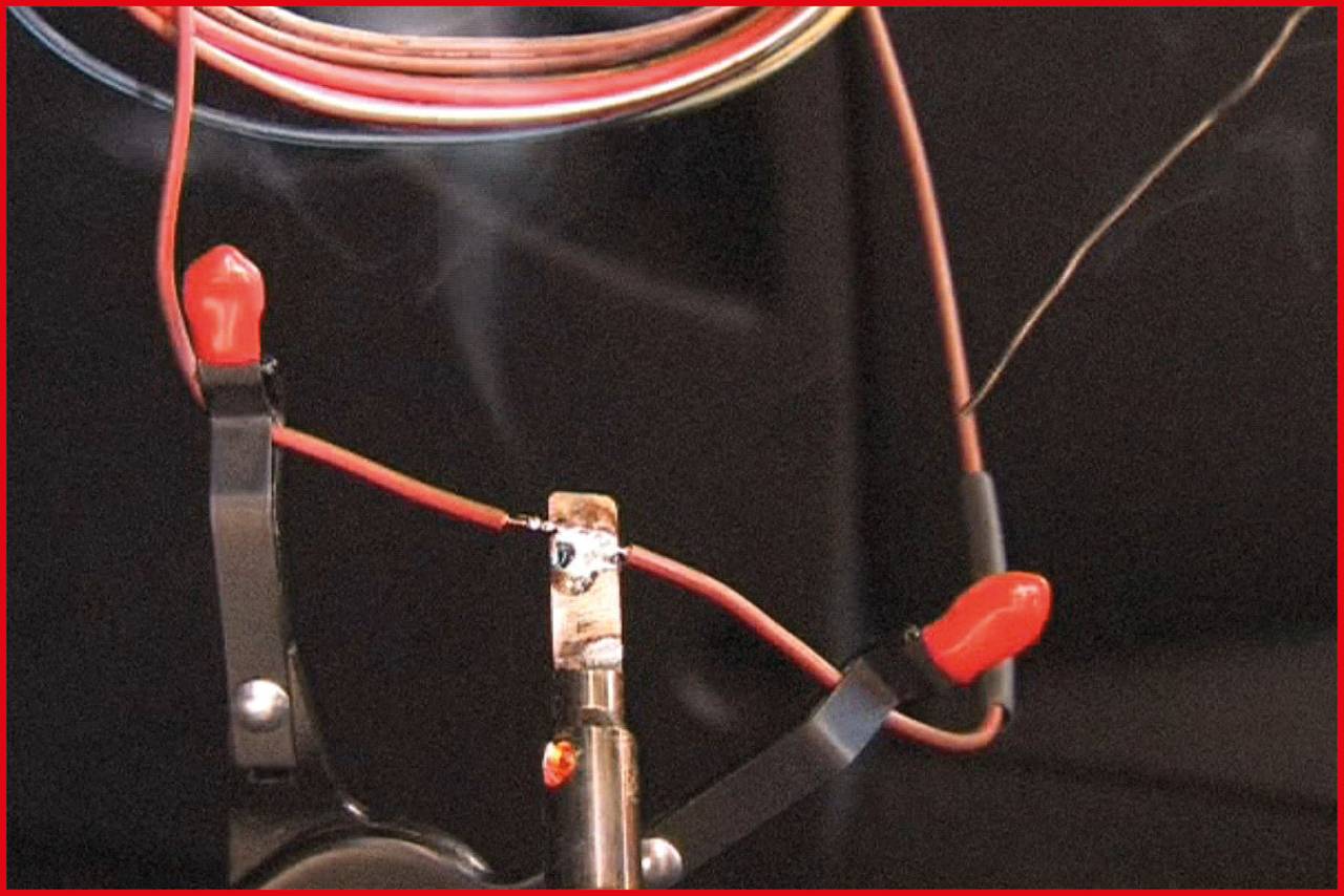 Le fer à souder touche le câble, produisant de la fumée. Les extrémités des câbles sont fixées avec des pinces rouges. Travail de câblage électrique en cours.