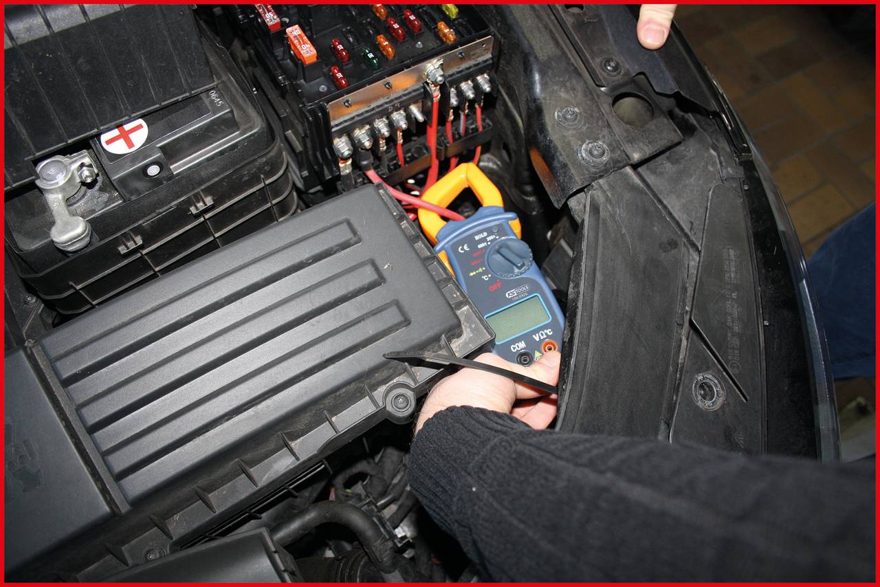 Une personne mesure la tension d'une batterie de voiture à l'aide d'un multimètre. La batterie est montée dans un boîtier noir.