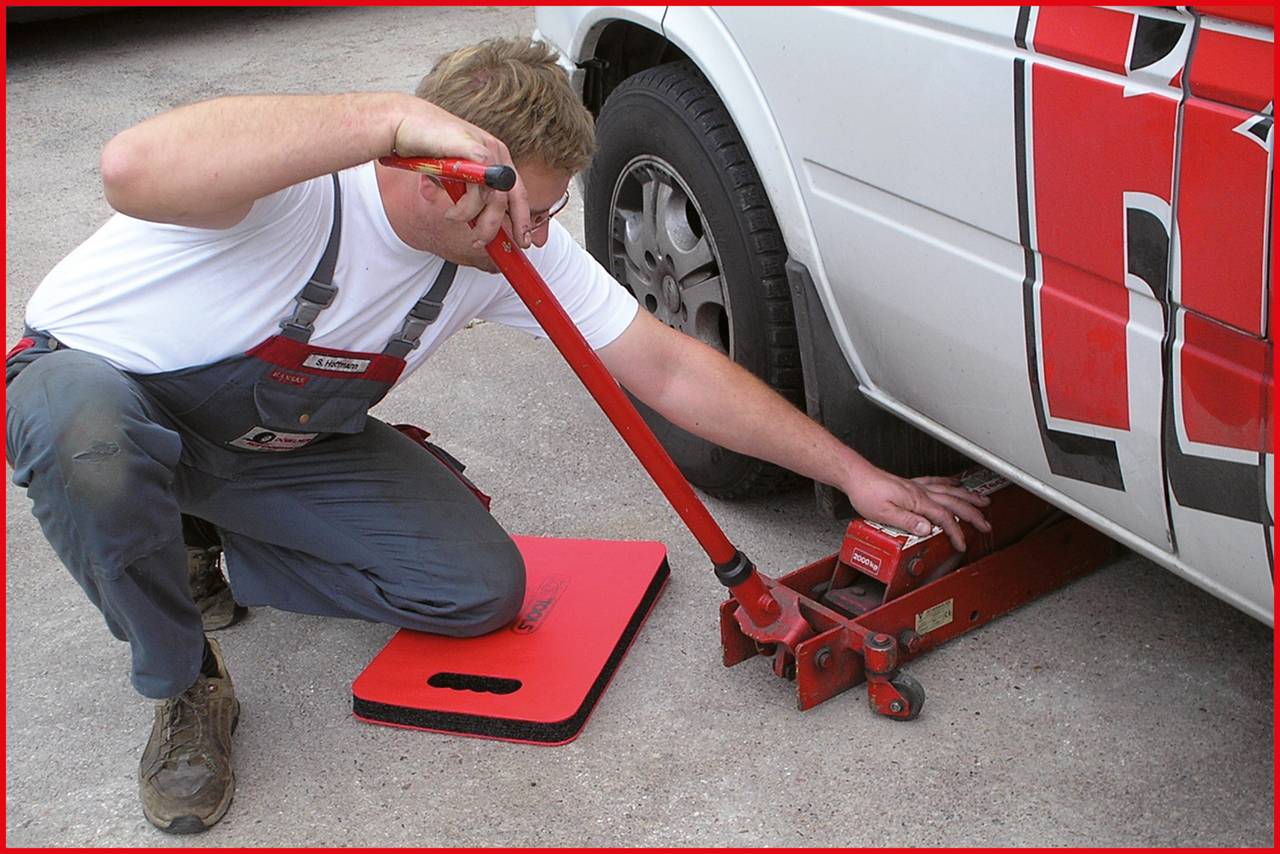 Un homme change un pneu, utilisant un cric pour soulever une voiture. Il porte des vêtements de travail et est à genoux sur le sol.