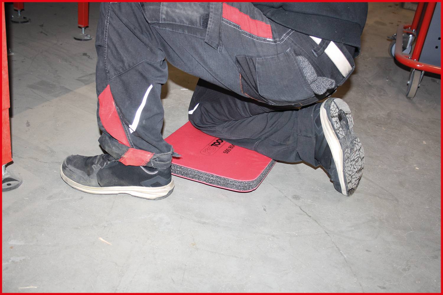 Une personne s'agenouille sur un tapis rouge sur un sol en béton, portant des chaussures de travail noires et un pantalon noir avec des bandes rouges.