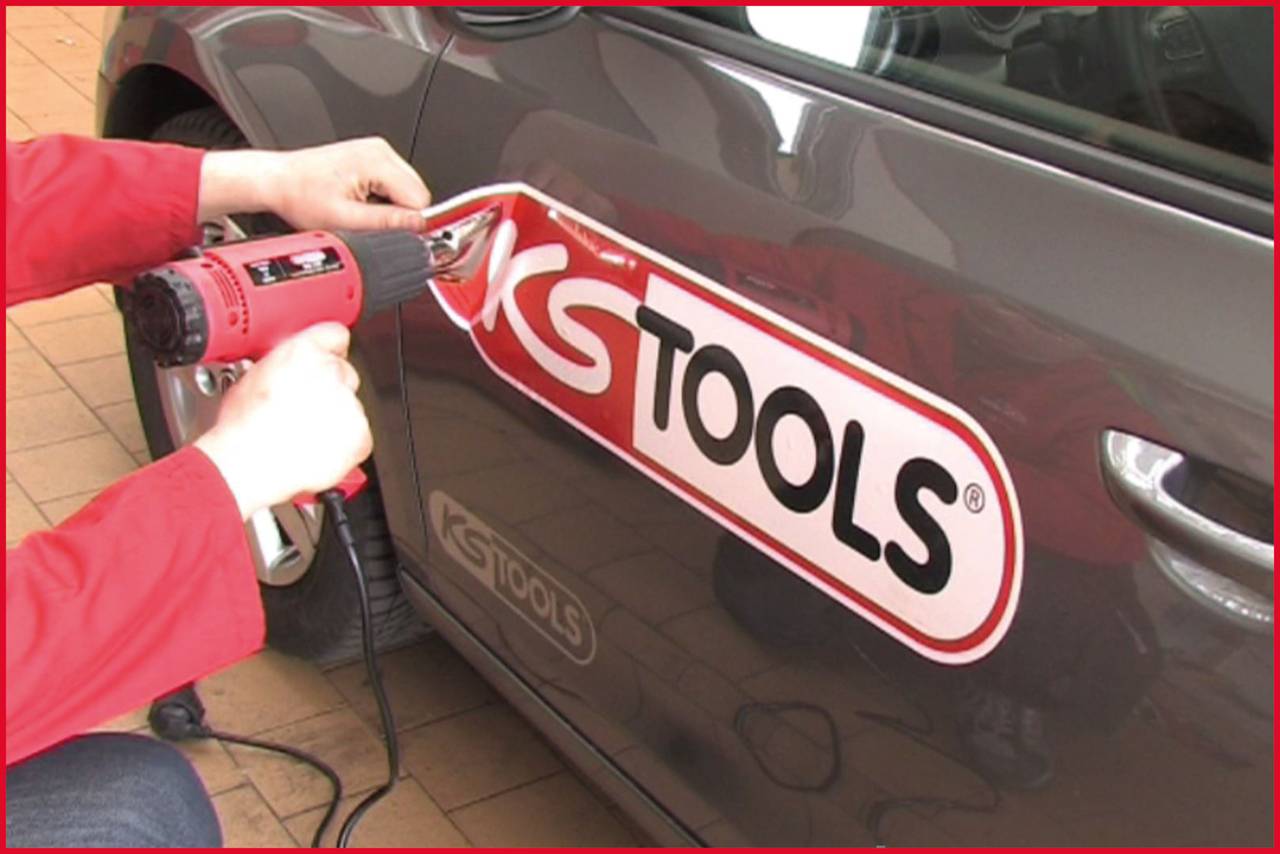 Une personne retire un grand autocollant de la portière d'un véhicule gris à l'aide d'un sèche-cheveux. L'autocollant porte l'inscription 'KS TOOLS'.
