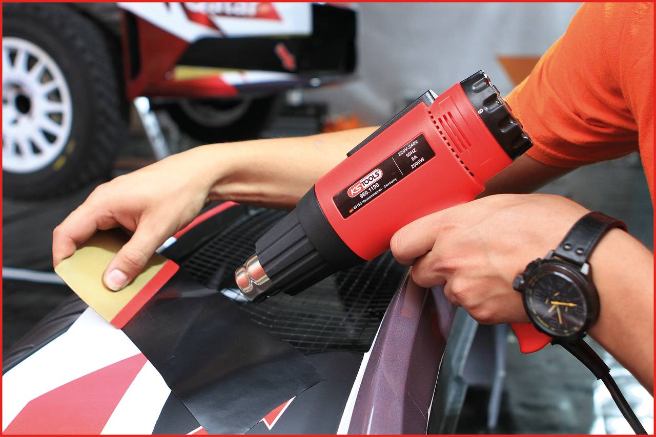 Une personne utilise un pistolet à air chaud pour appliquer un film vinyle sur une voiture ; gros plan sur les mains, le film et l'outil en premier plan.