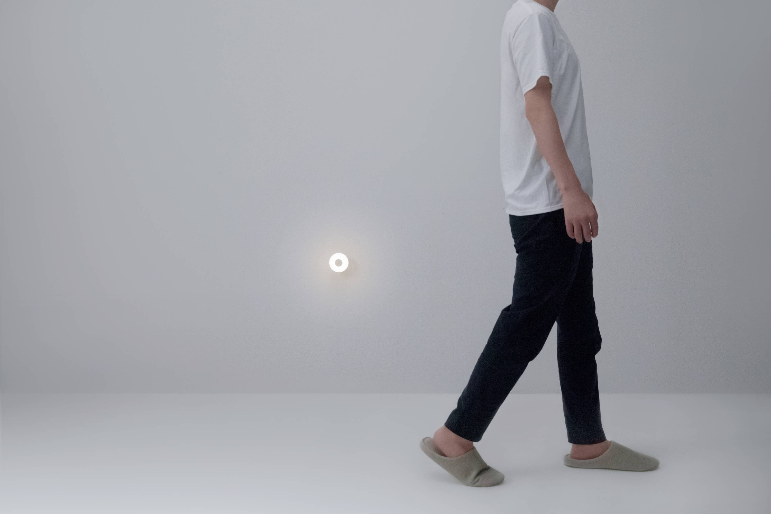 Une personne portant un t-shirt blanc et un pantalon noir marche vers la droite. À l'arrière-plan, on aperçoit une petite lumière ronde et lumineuse sur le mur.