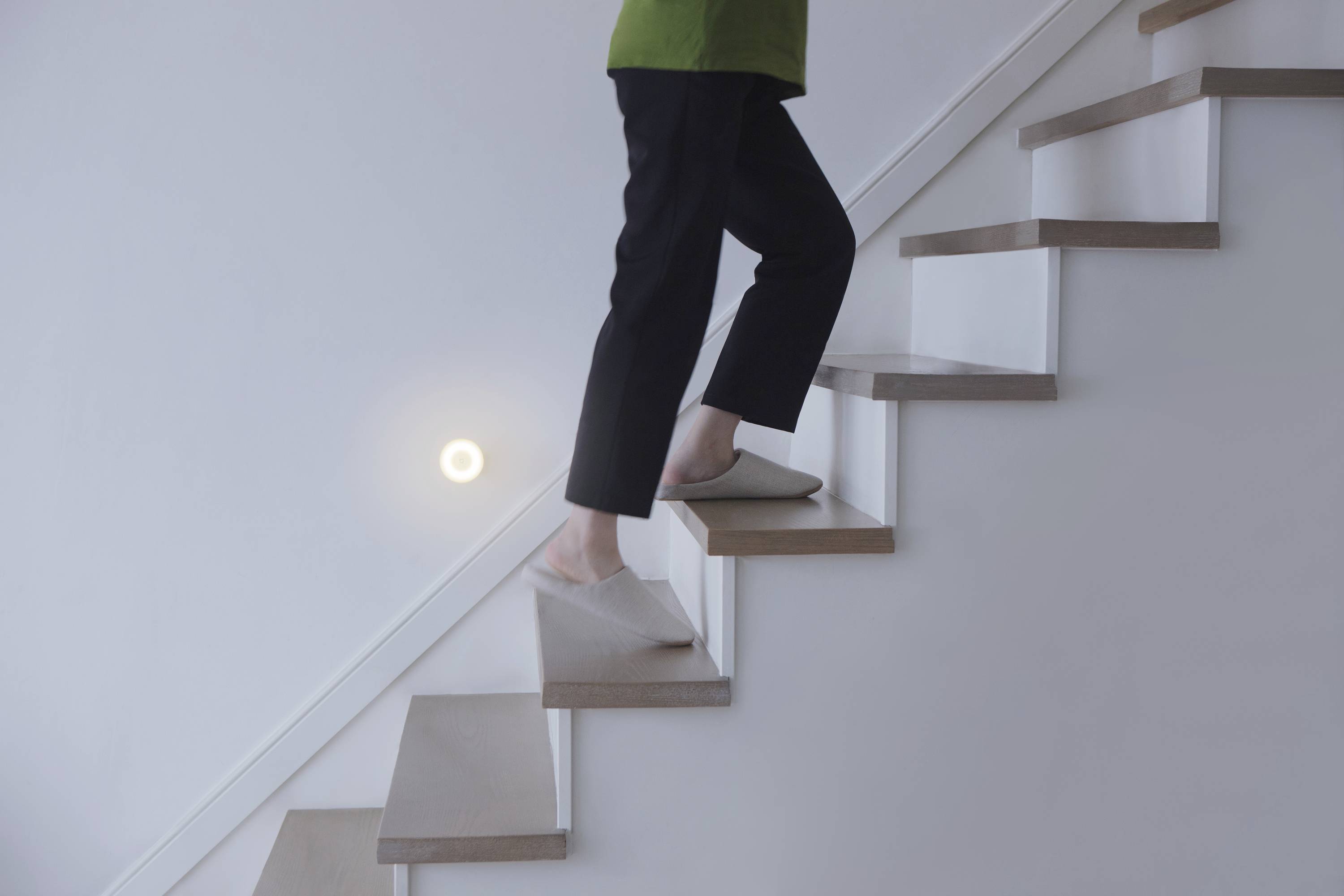 Une personne en pantoufles monte un escalier en bois, éclairée par un applique murale ronde.