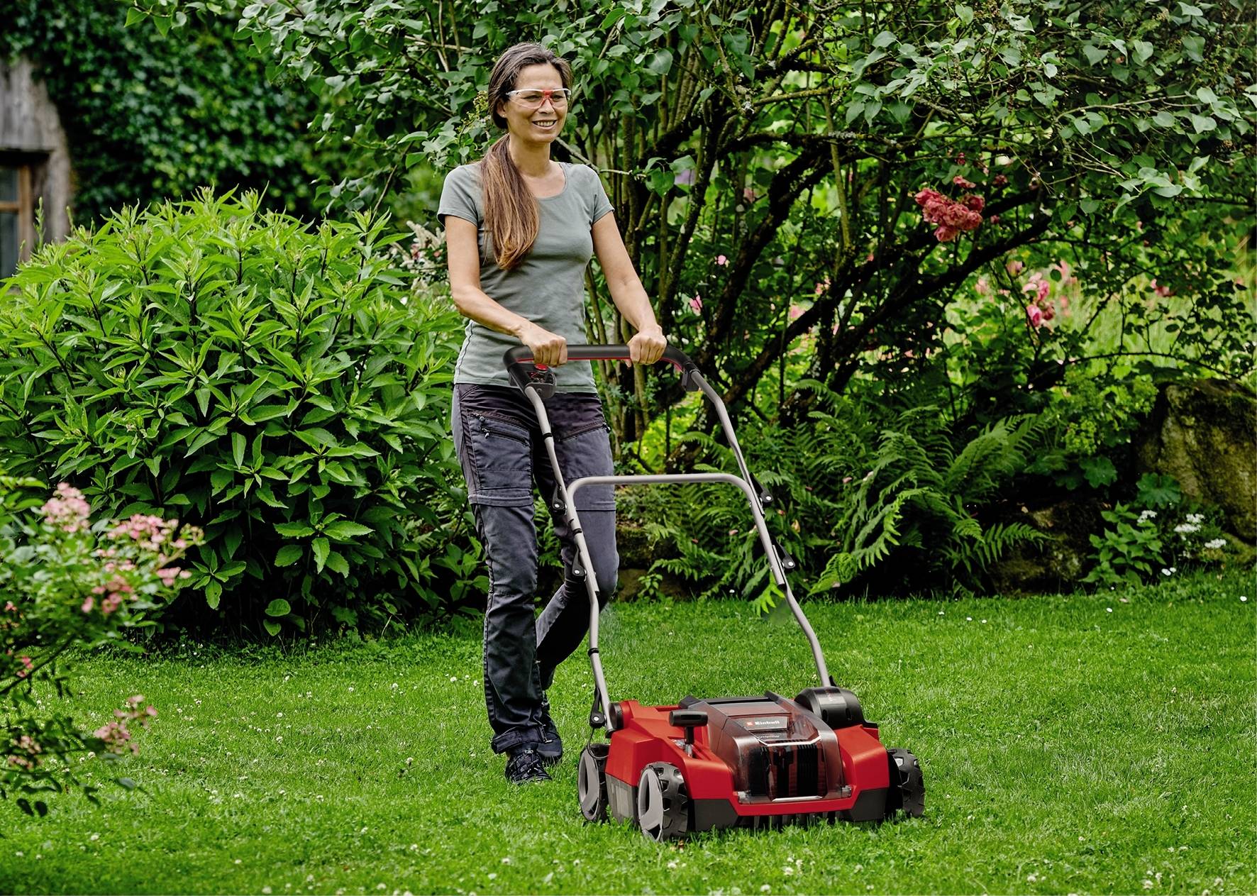 Une femme tond la pelouse avec une tondeuse rouge dans un jardin, entourée de plantes et d'arbres verts.
