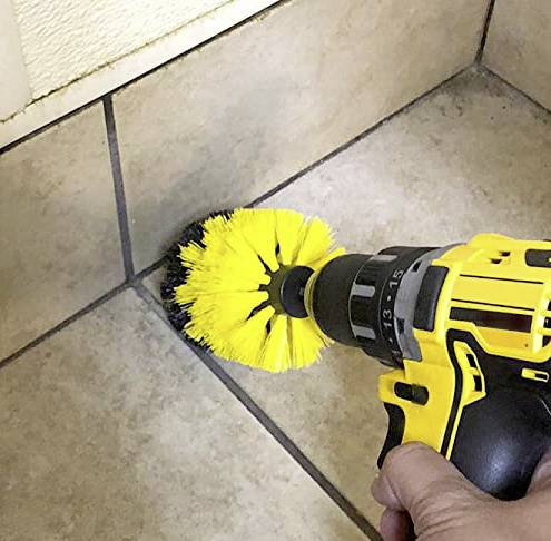 Une main tient un tournevis sans fil jaune équipé d'une brosse rotative qui nettoie les joints de carrelage. Des carreaux sont visibles en arrière-plan.