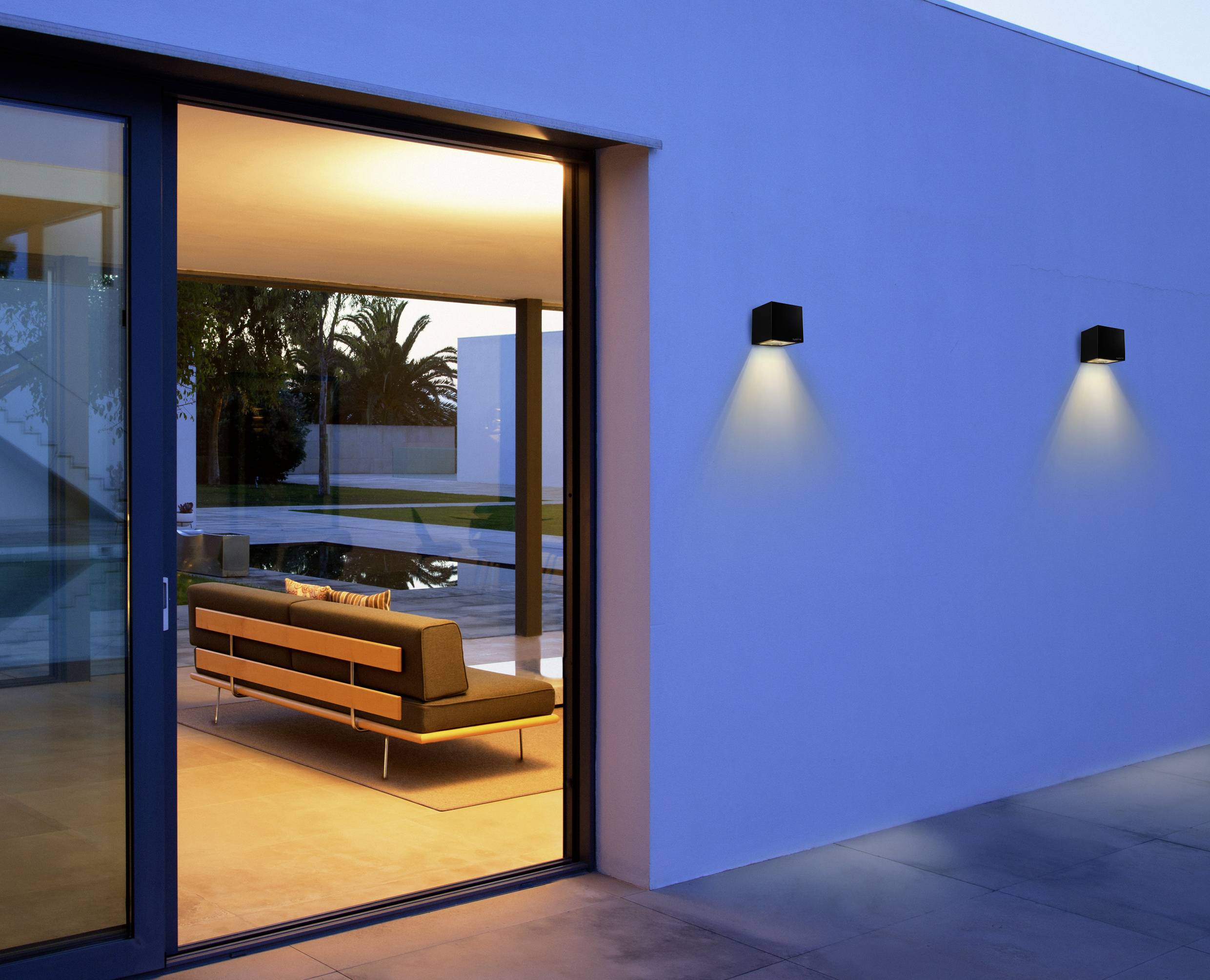 Un salon moderne avec un intérieur éclairé et une grande fenêtre offrant une vue sur une terrasse avec une piscine et des palmiers.