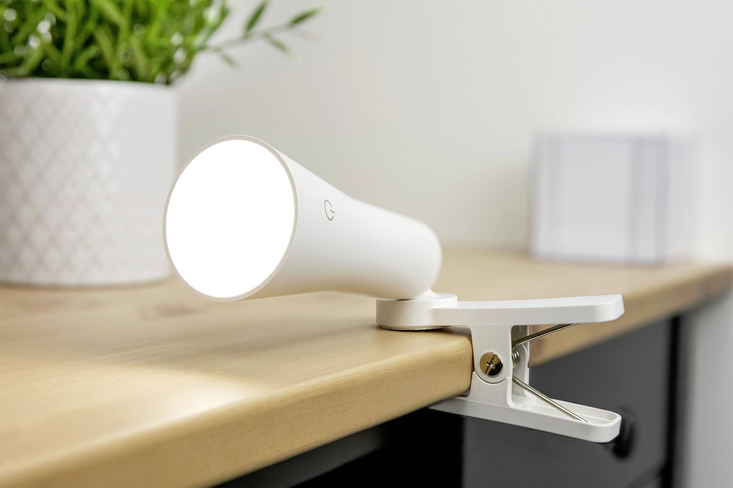 Lampe LED blanche à pince, fixée au bord d'une table, brillant intensément. En arrière-plan, flou, une plante et un objet cubique.