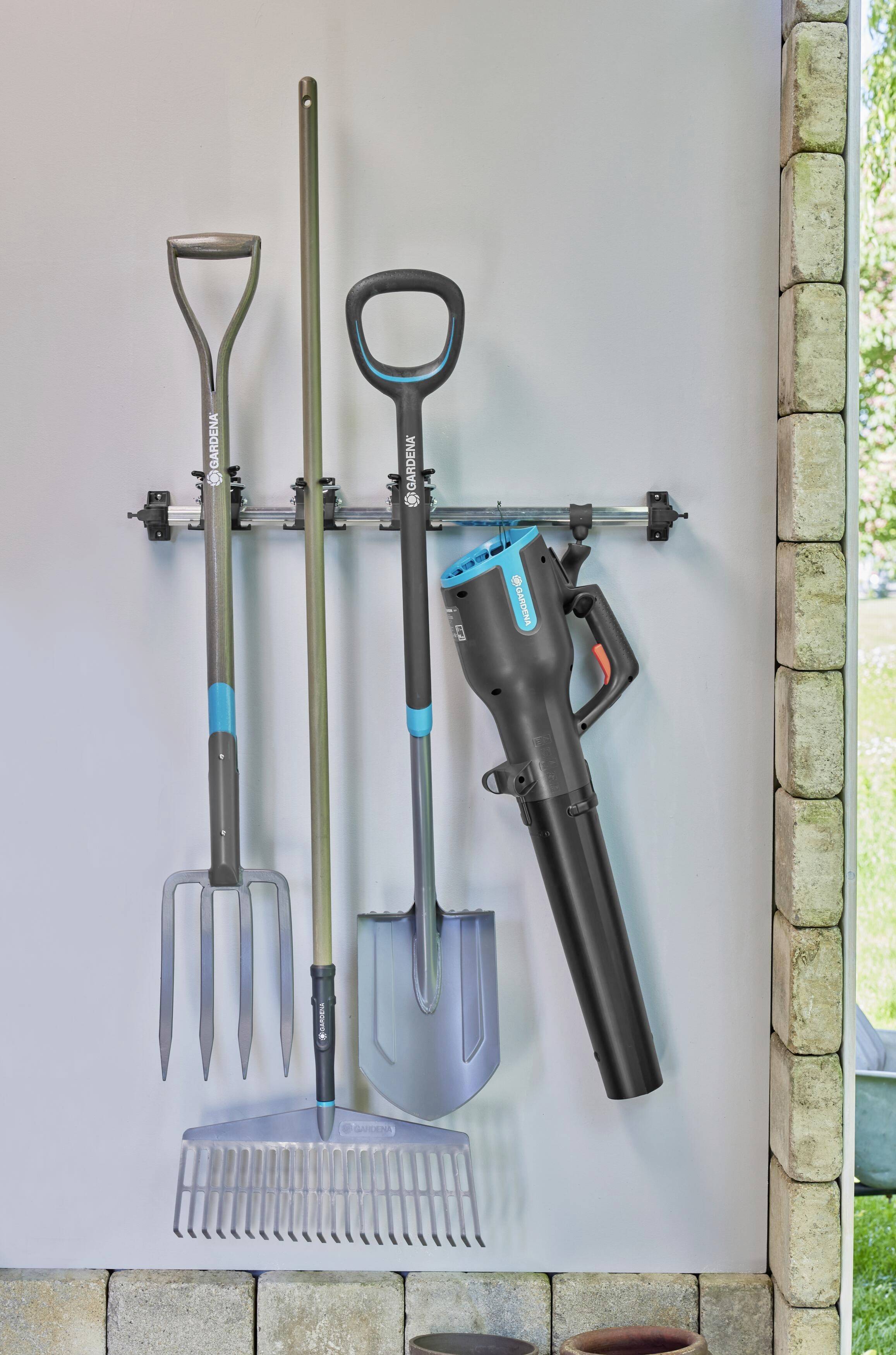 Des outils de jardin sont suspendus à un support mural. On peut y voir une fourche à fumier, un râteau, une pelle et un souffleur de feuilles.