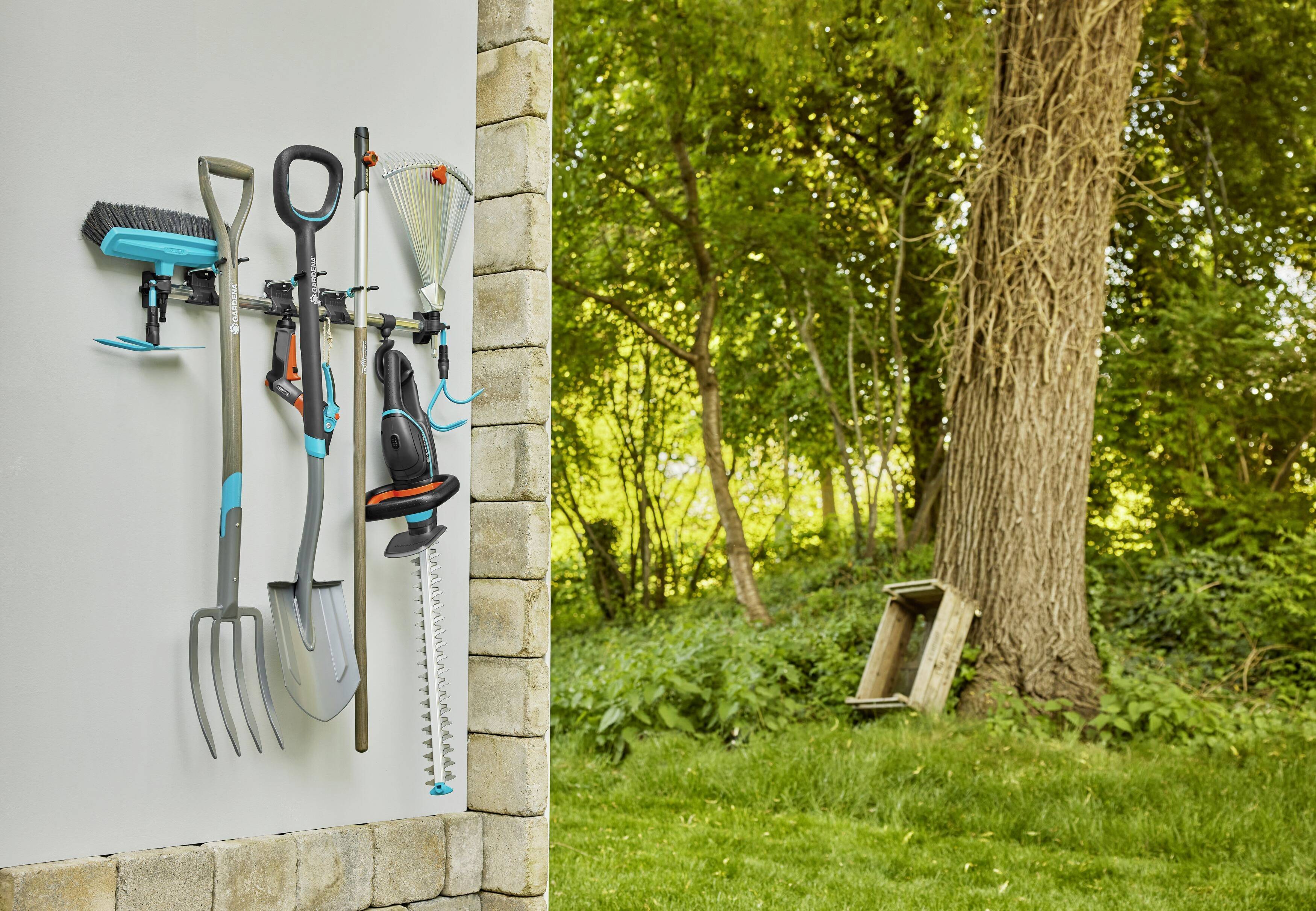 Des outils de jardin comme un balai, un râteau et un taille-haie sont suspendus de manière ordonnée à un support mural. En arrière-plan, un arbre et une pelouse verte.
