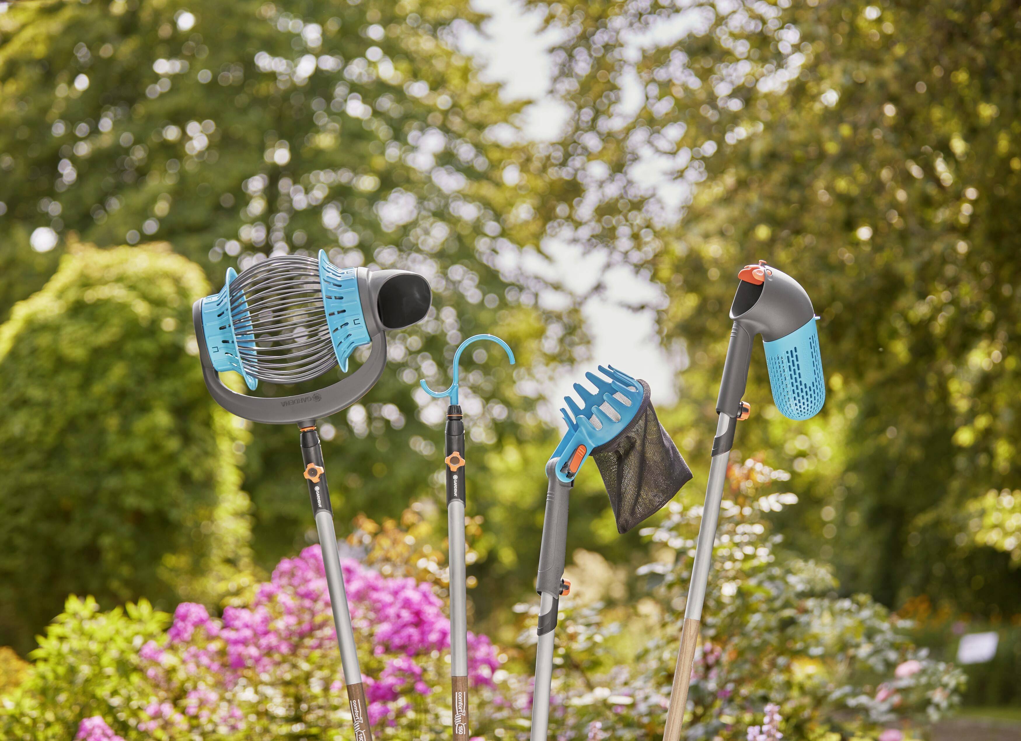 Outils de jardin aux poignées bleues et noires : un balai à feuilles, un râteau à main et une pelle, dressés verticalement dans un jardin avec des buissons en fleurs.