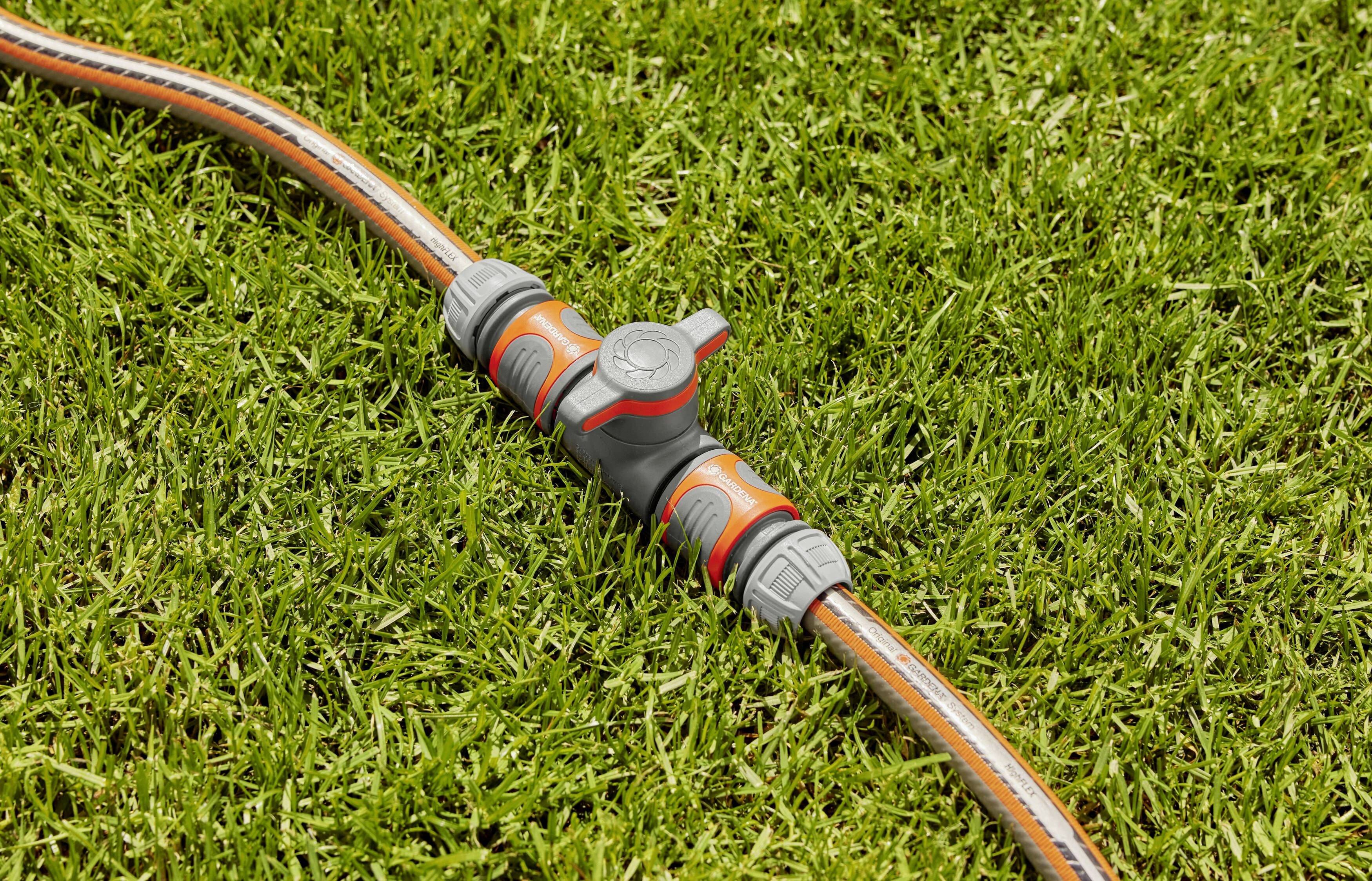 Un tuyau d'arrosage est posé sur de l'herbe verte, relié par une valve grise en forme de T.