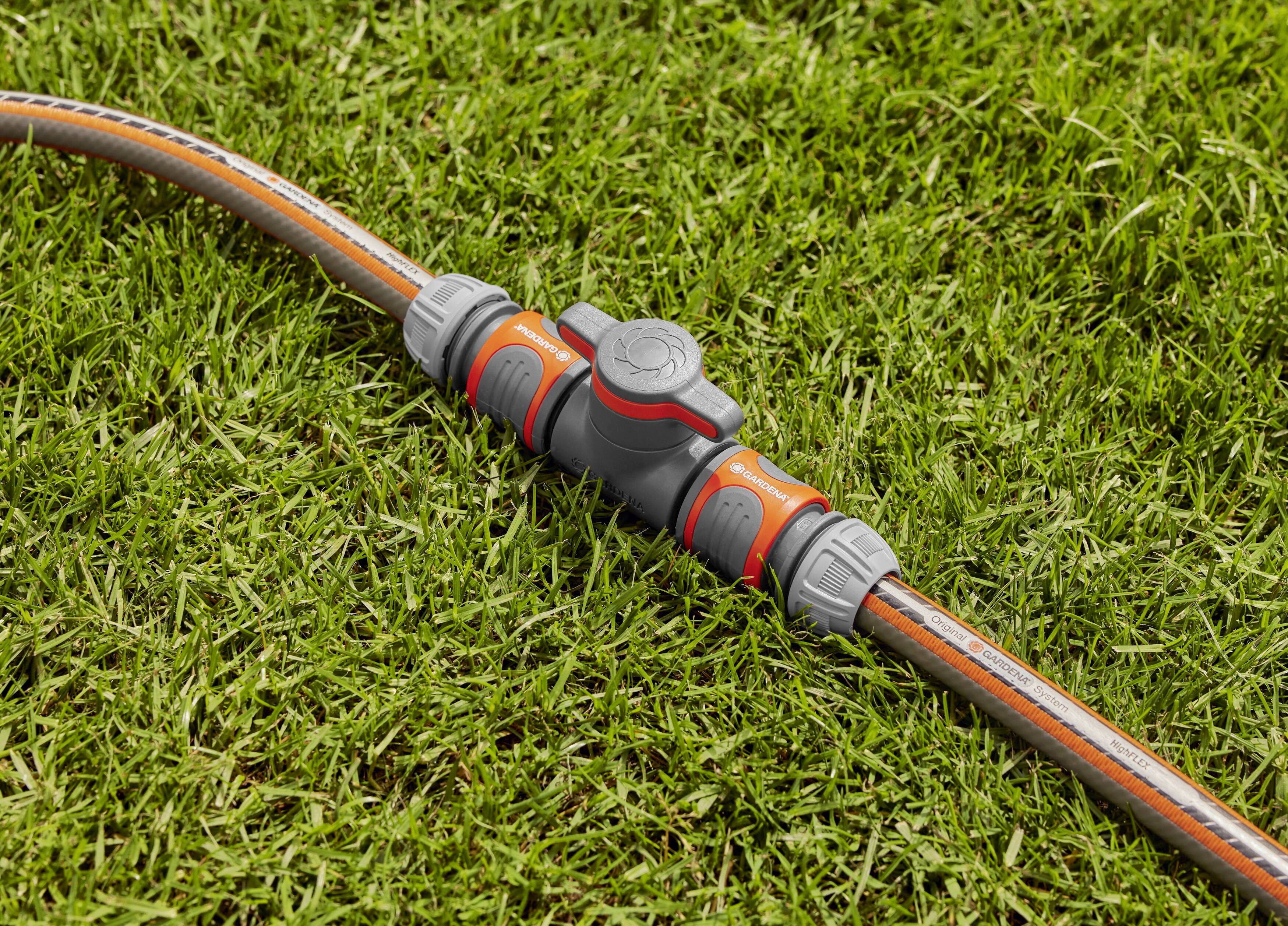 Un connecteur de tuyau d'arrosage gris avec un levier rouge repose sur une pelouse verte. Le connecteur relie deux tuyaux entre eux.