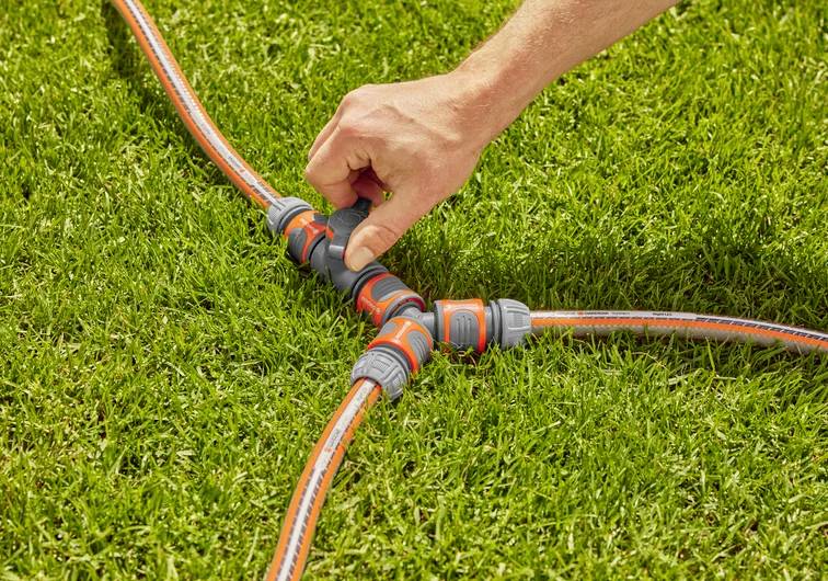 Une main relie trois tuyaux d'arrosage à l'aide d'un répartiteur sur de l'herbe verte, illustrant une méthode pour distribuer l'eau dans différentes zones.