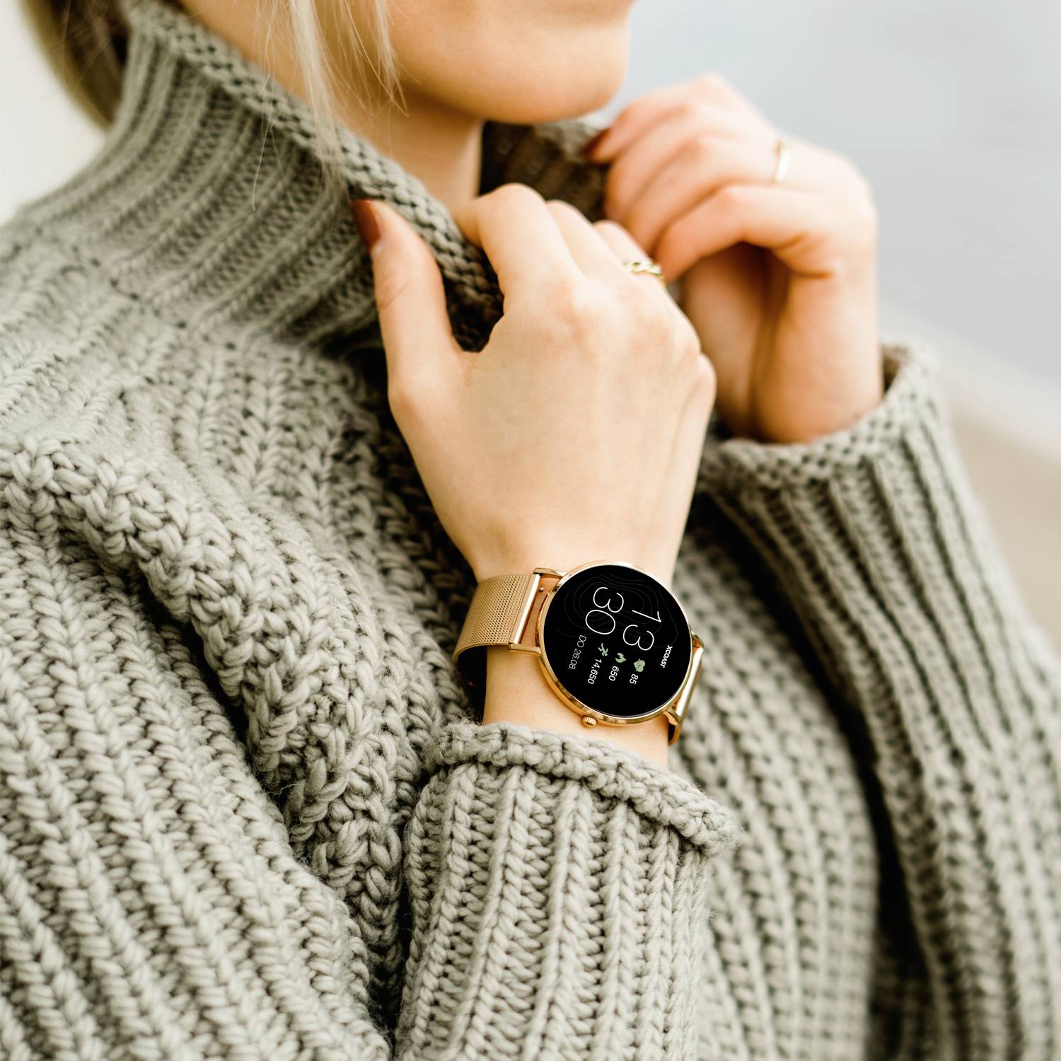 Une personne portant un pull en tricot gris porte une montre intelligente au poignet, affichant l'heure, la fréquence cardiaque et le nombre de pas.