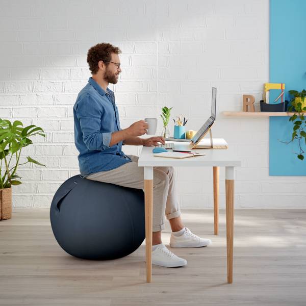 Un homme est assis sur un ballon de gymnastique à un bureau, travaillant sur un ordinateur portable. Bureau moderne et lumineux avec des plantes.