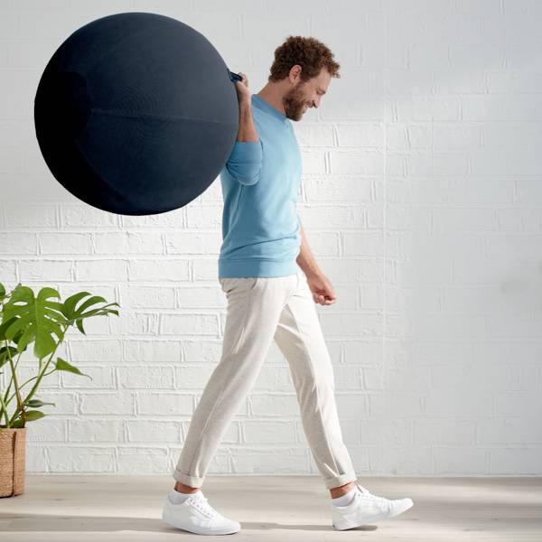Un homme en tenue décontractée fait un équilibre avec un ballon de gymnastique noir sur son dos. Une plante est visible en arrière-plan.