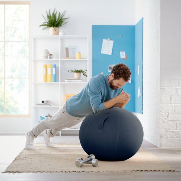 Un homme en tenue de sport s'appuie sur un ballon de fitness dans le salon. Des étagères et un tableau d'affichage sont visibles en arrière-plan.