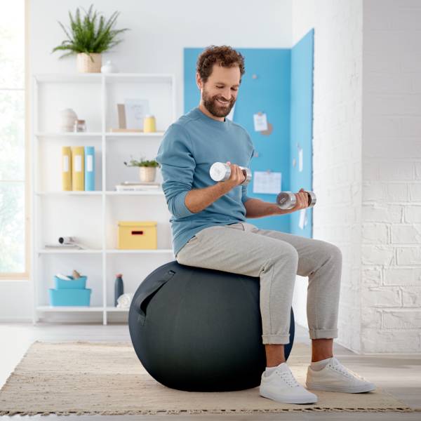 Un homme s'entraîne dans le salon avec des haltères, assis sur un ballon de fitness, portant des vêtements de sport confortables. En arrière-plan, une étagère avec des plantes.