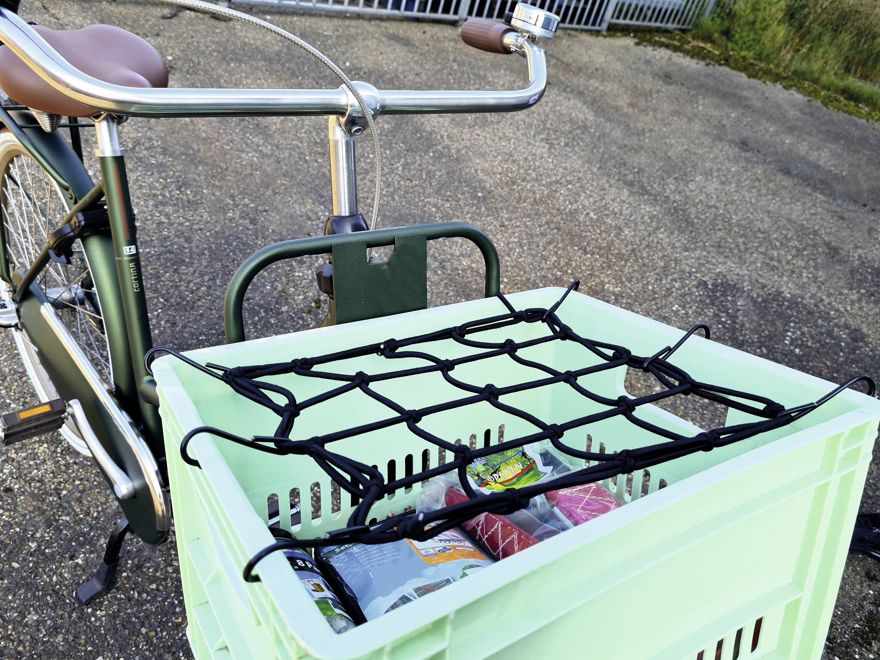 Vélo vert avec guidon en bois et panier, sécurisé par un filet de portage. Le panier contient des courses. En arrière-plan, de l'asphalte.