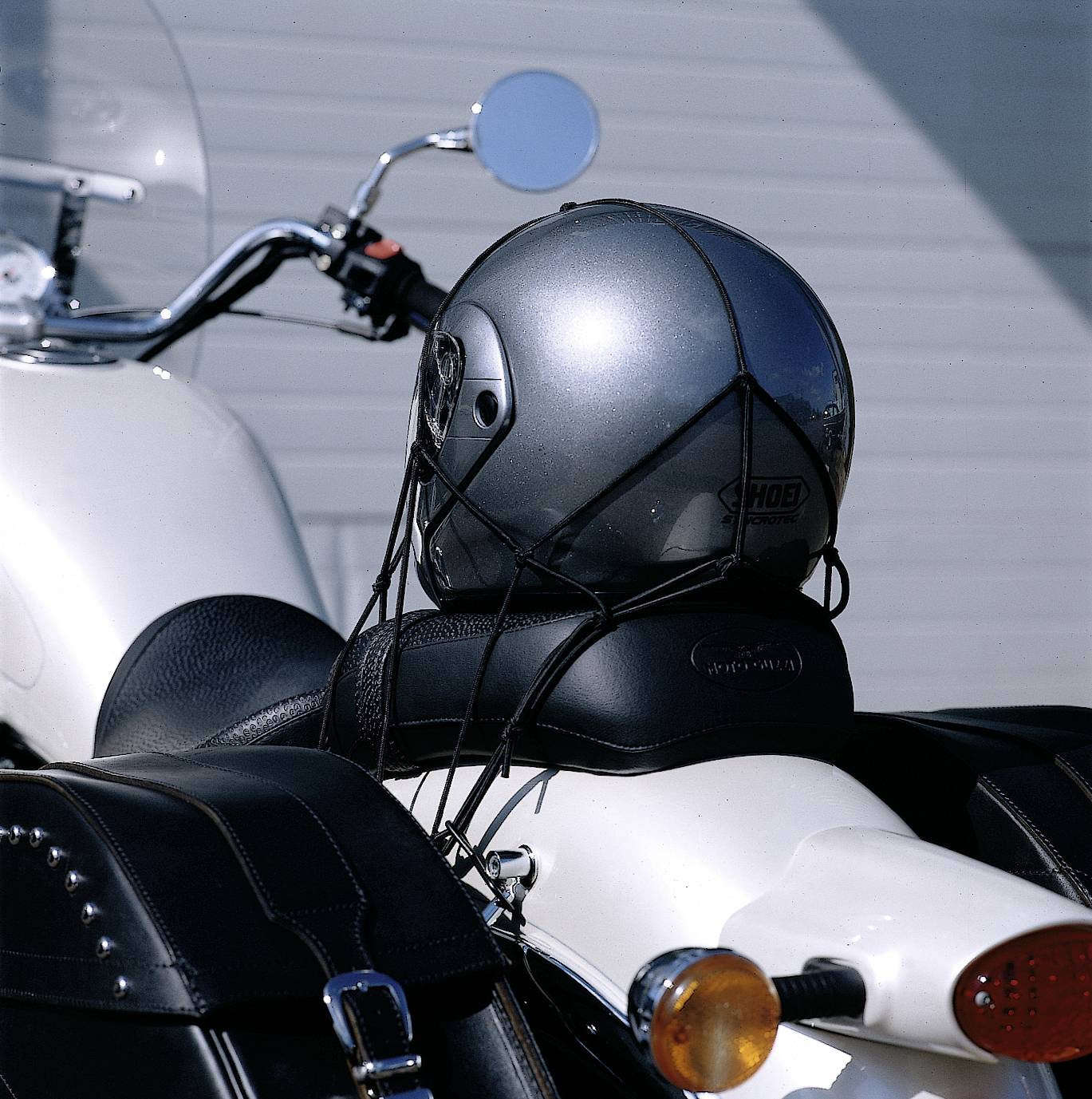 Un casque est attaché avec des sangles sur le siège d'une moto blanche. À gauche se trouvent le guidon et le rétroviseur. En arrière-plan, un mur.