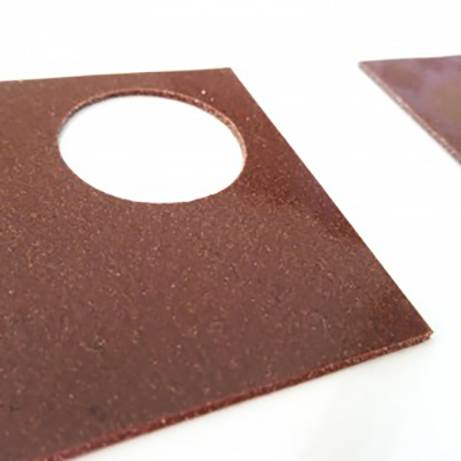 Plaque métallique carrée brune avec un trou circulaire découpé au centre, surface légèrement rugueuse.