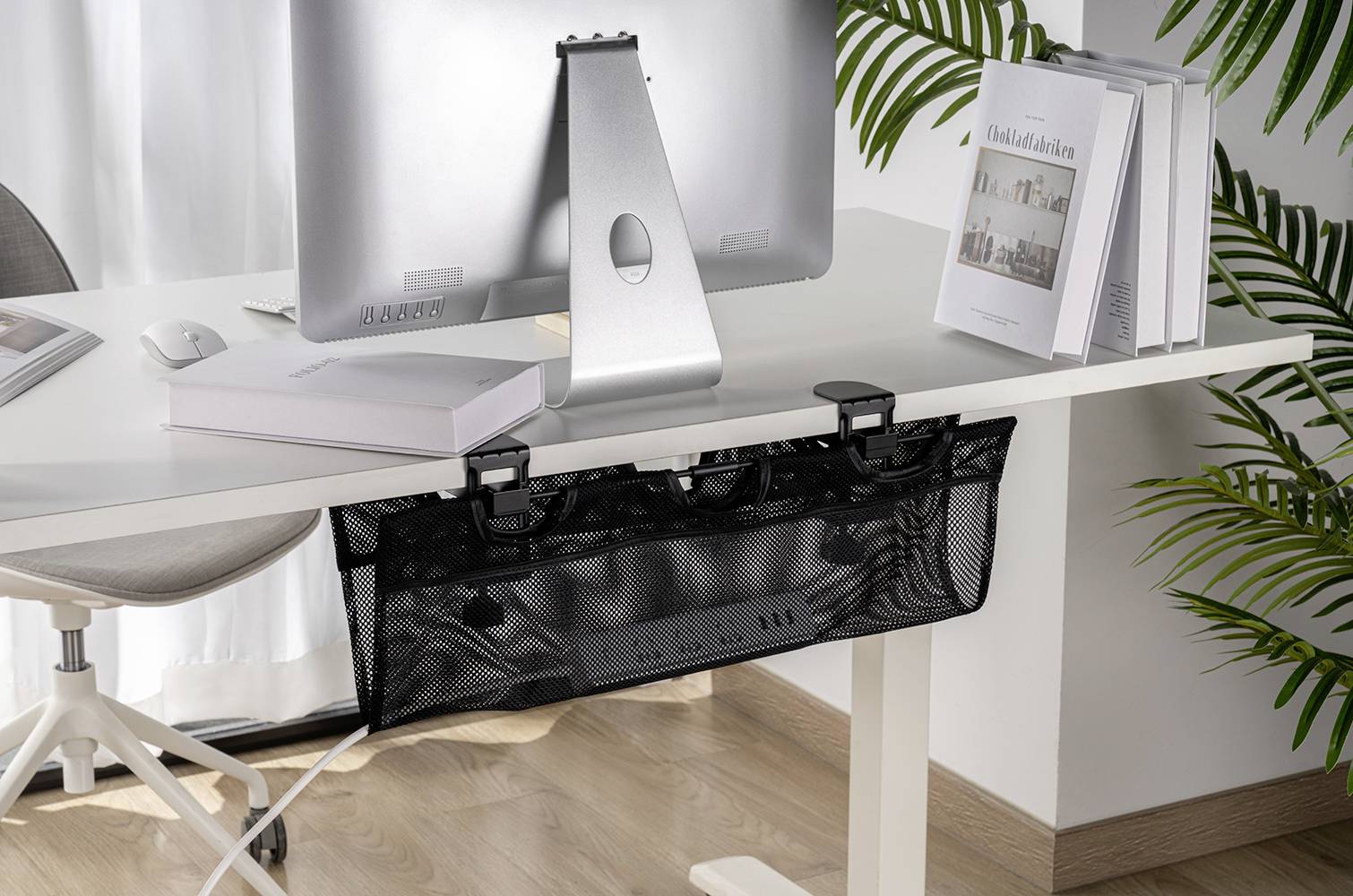 Un bureau avec un ordinateur et un panier de gestion des câbles en dessous. Des livres sont posés sur la table, et des plantes sont visibles en arrière-plan.