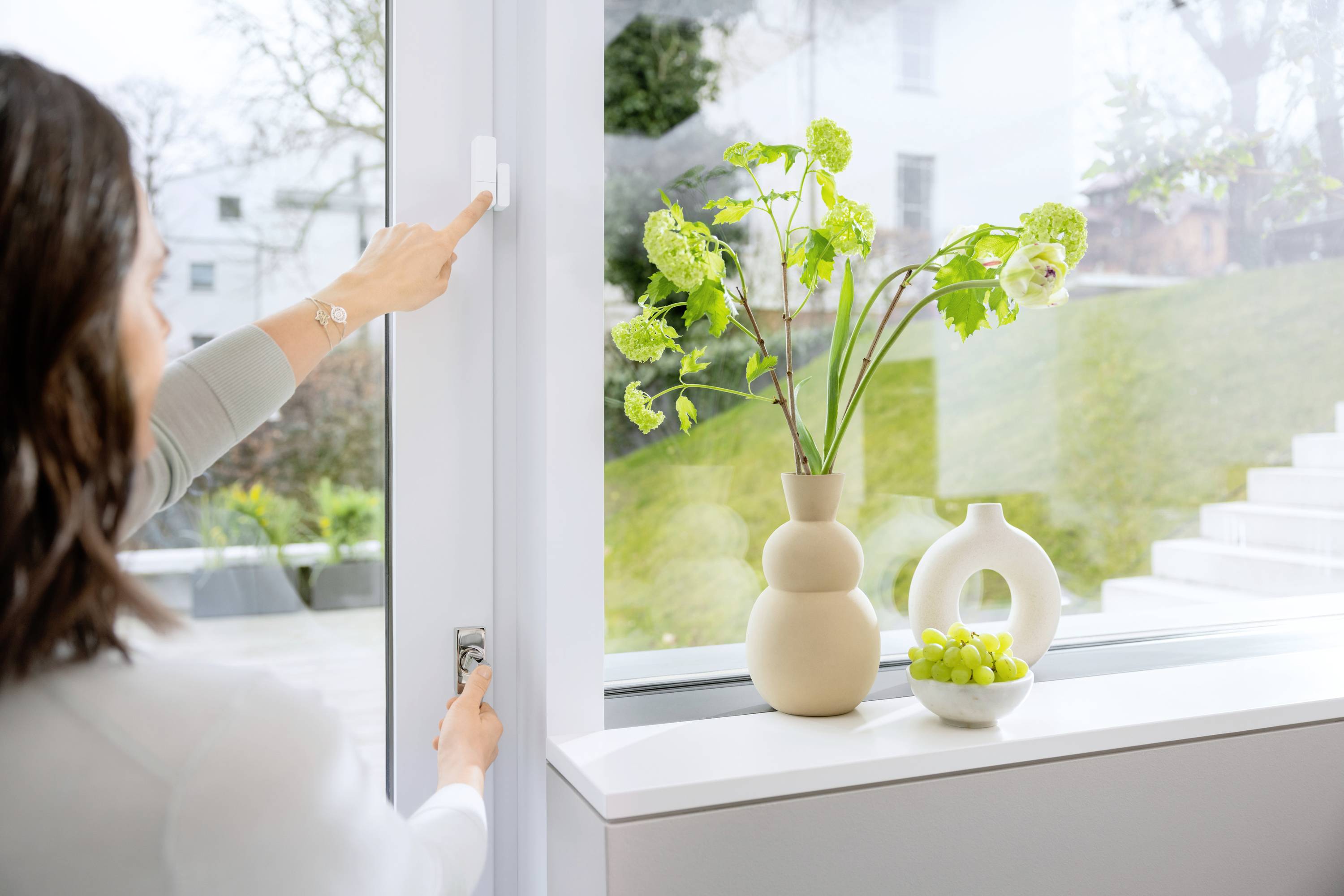 Une personne ouvre une fenêtre ; sur le rebord de la fenêtre se trouvent un vase avec des fleurs et un bol rempli de fruits verts.