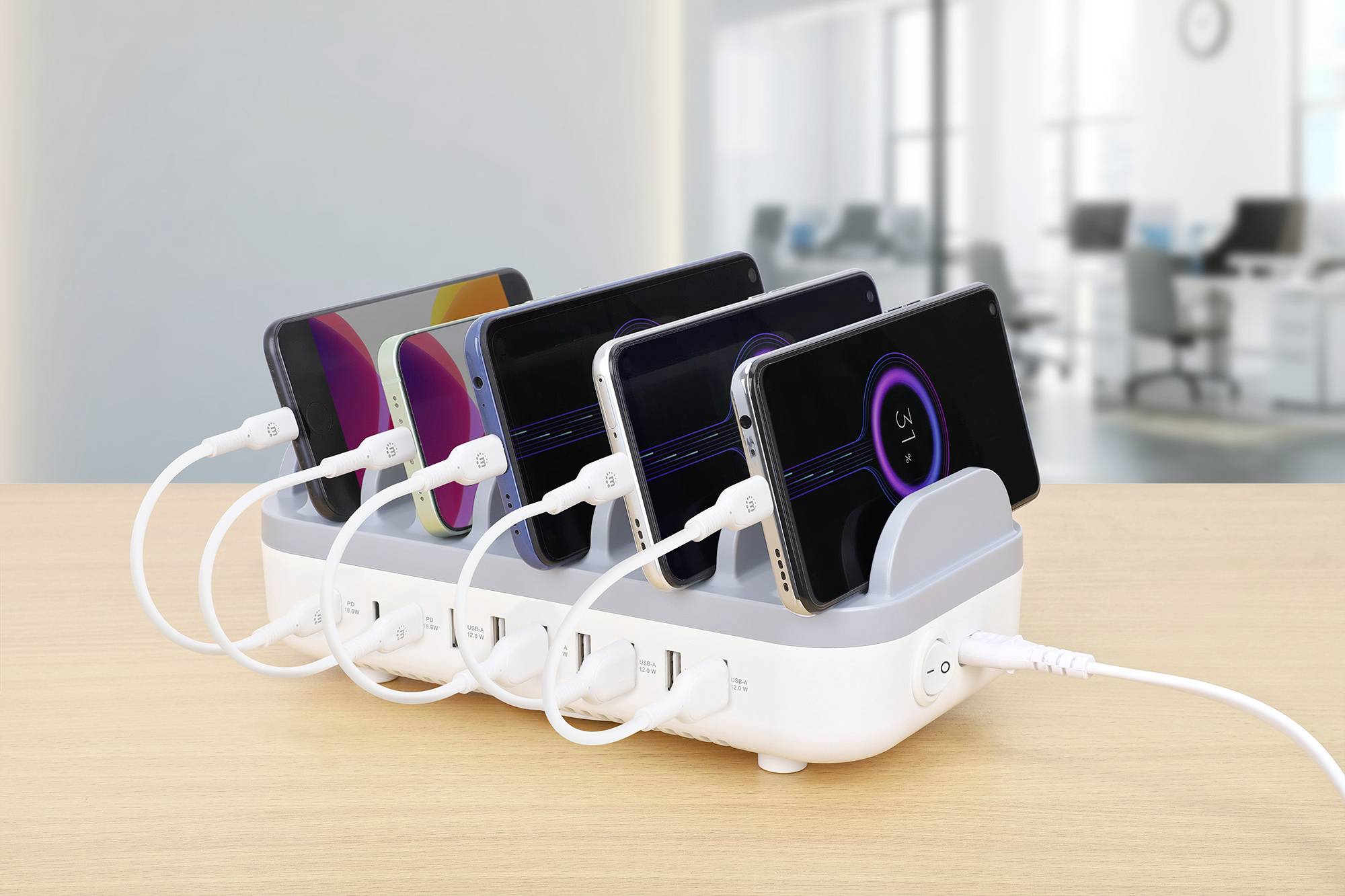 Plusieurs smartphones sont en cours de chargement simultanément sur une station de recharge. En arrière-plan, on aperçoit un bureau flou.
