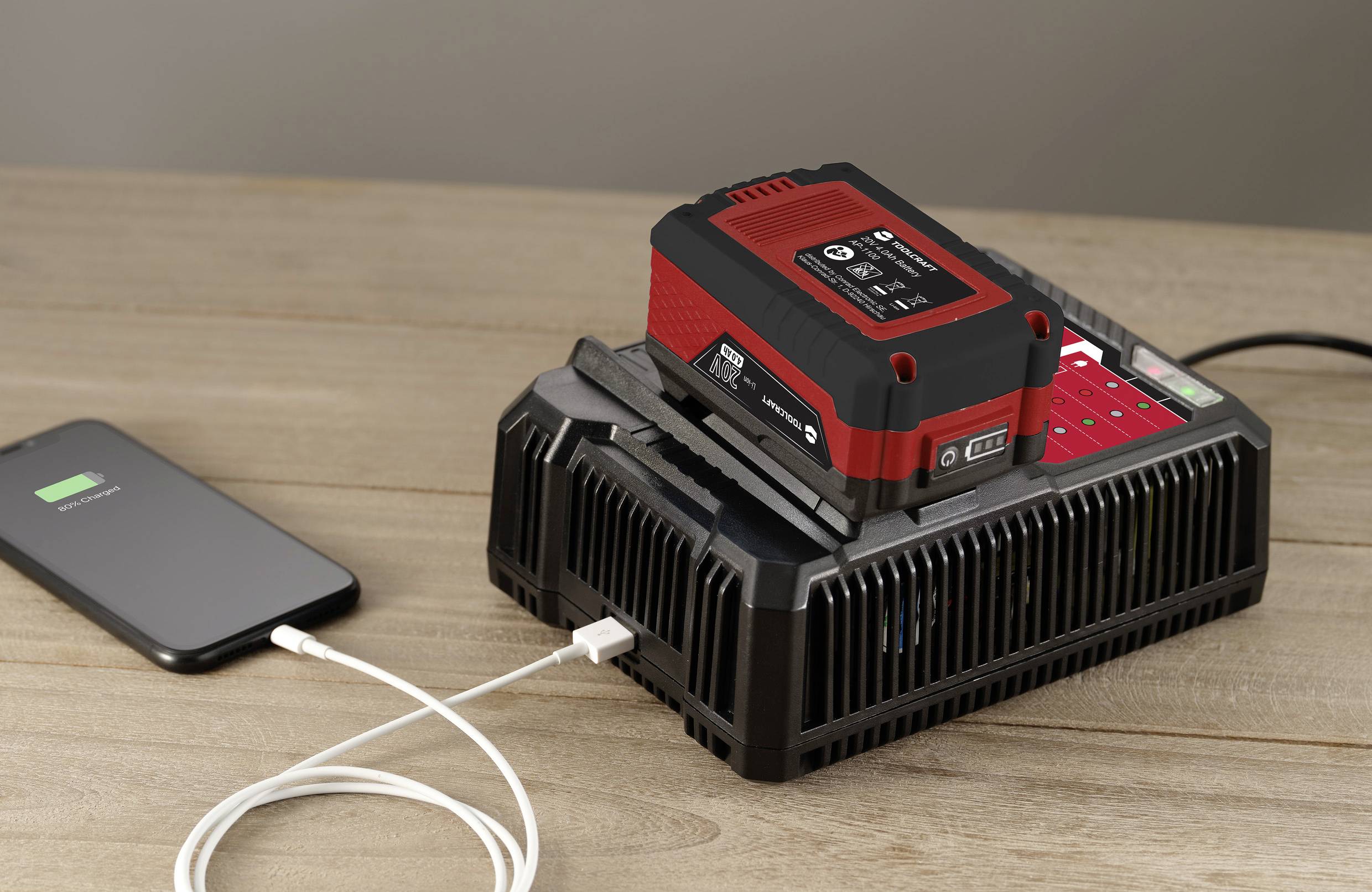 Un chargeur de batterie charge une batterie rouge. Un smartphone avec un indicateur de charge est connecté au chargeur via un câble.