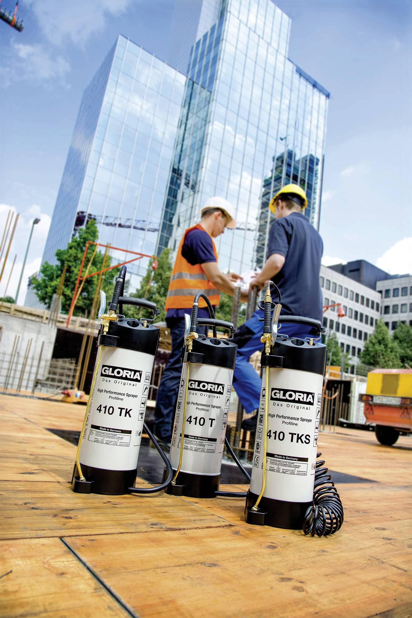 Deux ouvriers du bâtiment discutent des plans sur un chantier ; au premier plan, trois extincteurs sont disposés ; à l'arrière-plan, des immeubles de bureaux modernes sont visibles.
