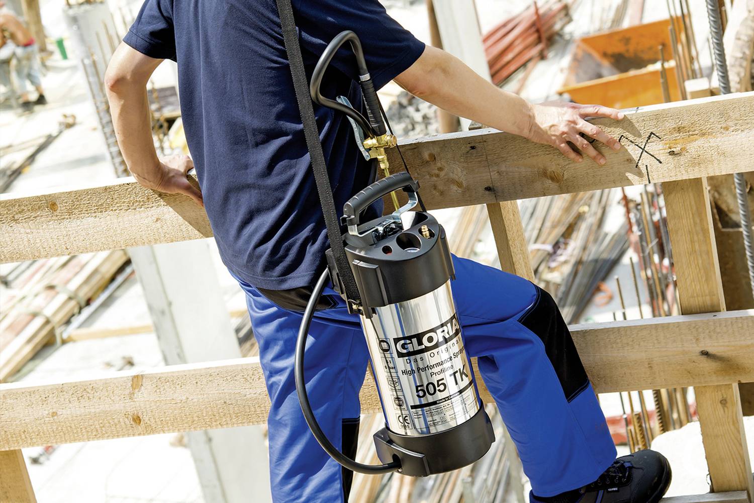 Personne sur un chantier de construction portant un réservoir métallique avec un tuyau sur le dos, touchant une rambarde en bois, avec des échafaudages en arrière-plan.