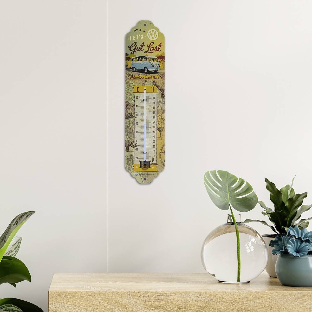 Thermomètre mural au design VW avec l'inscription 'Let's Get Lost' placé au-dessus d'une étagère. Des plantes à gauche et à droite complètent la décoration.
