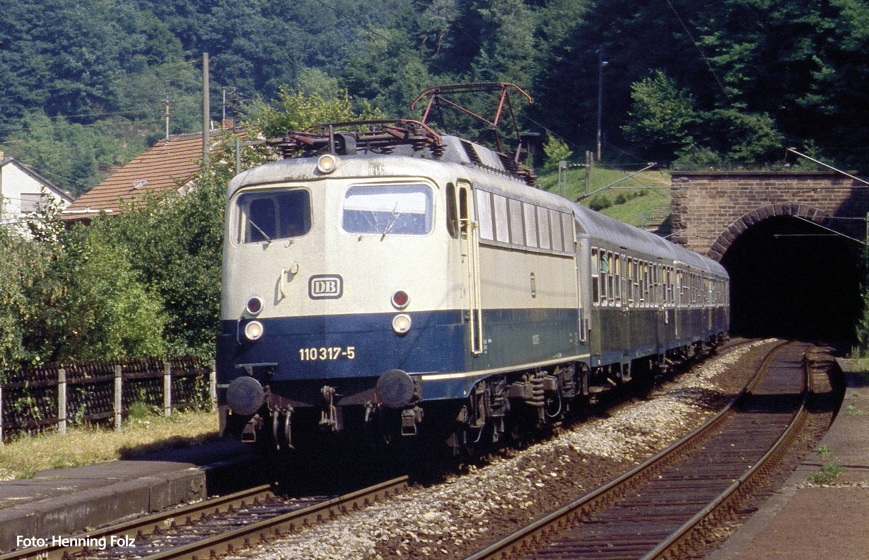 Un train bleu et blanc de la Deutsche Bahn sort d'un tunnel. Des forêts et des maisons sont visibles en arrière-plan.