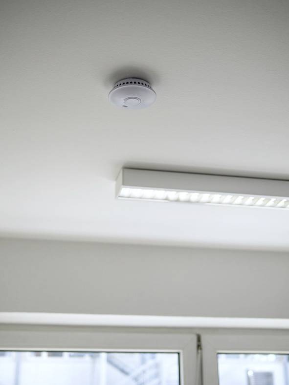 Un détecteur de fumée est installé sur un plafond blanc près d'un tube fluorescent, dispositif de sécurité important dans la pièce.